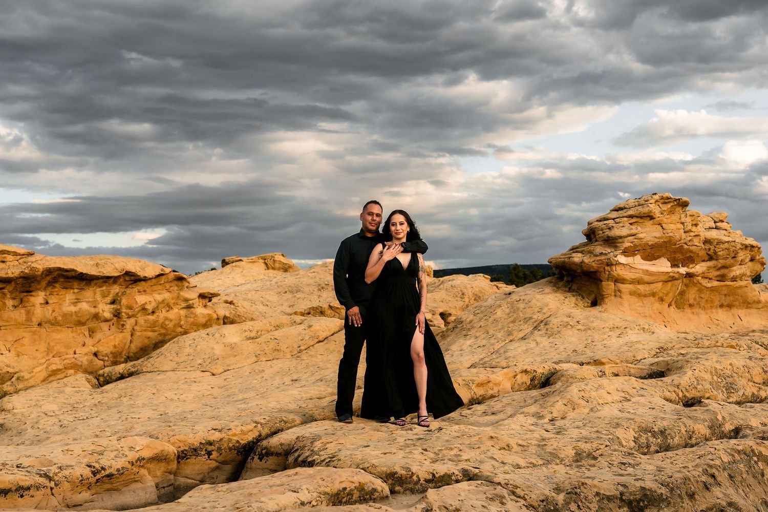 Couple in black outfits posing on golden sandstone rocks under dramatic cloudy sky at sunset.