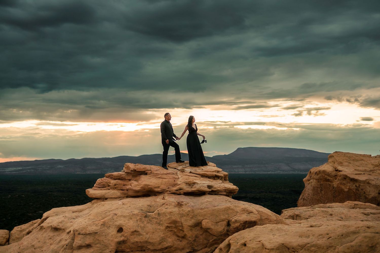 Couple holding hands on rocky summit at sunset with sweeping valley views and dramatic cloudy sky.