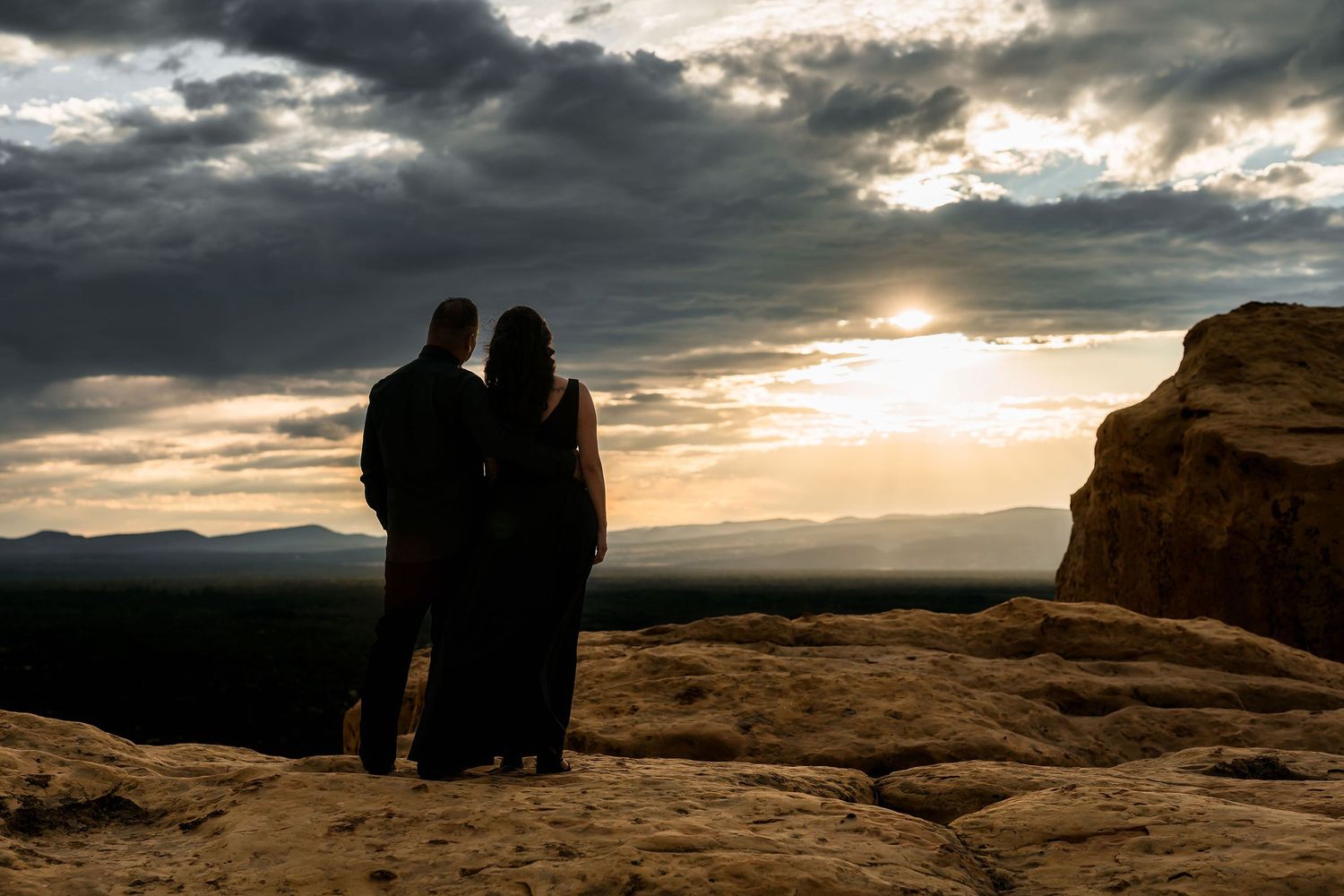 Couple silhouetted against a dramatic sunset sky on a rocky desert cliff overlooking a vast landscape.