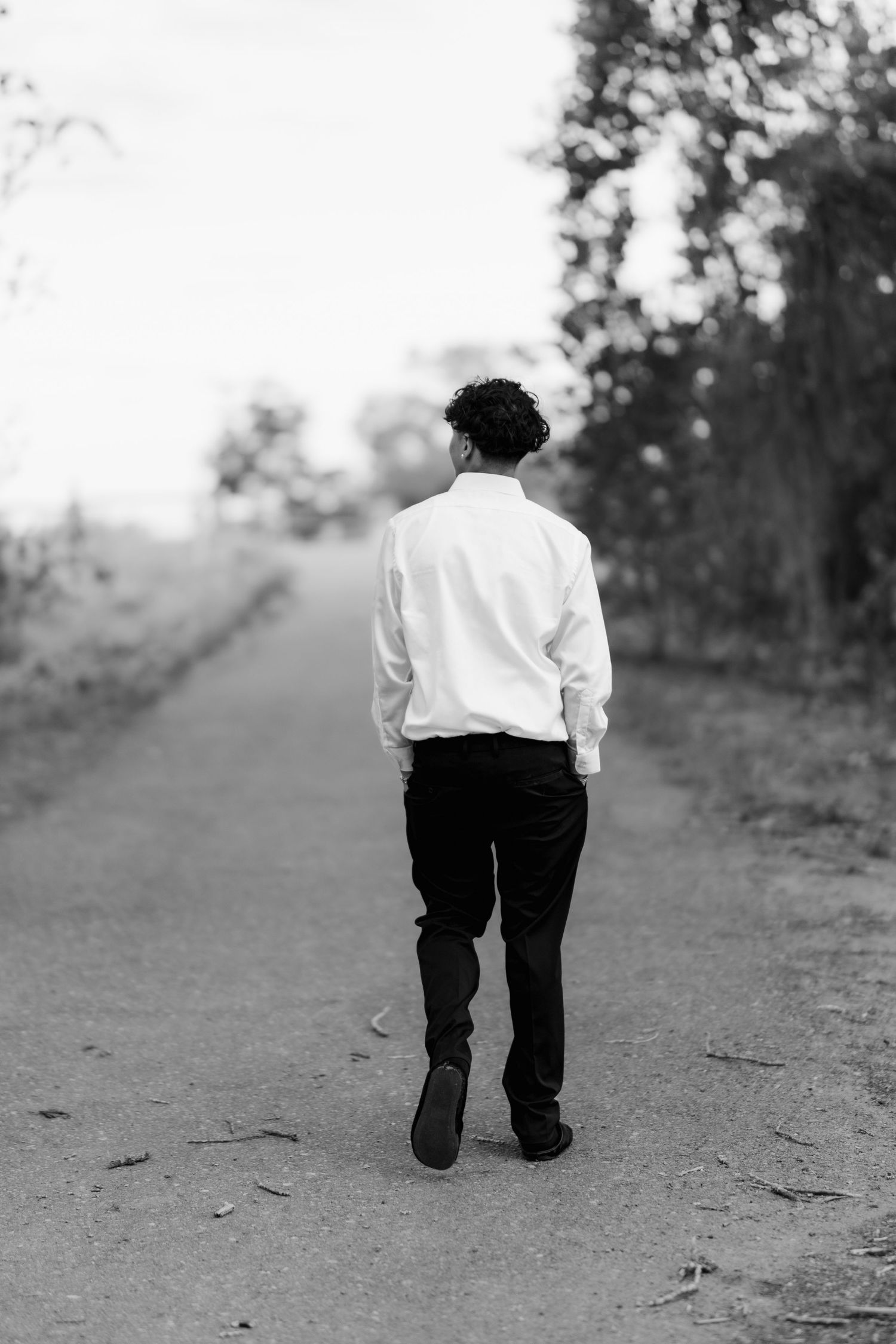 A monochrome photo of someone in a white shirt and dark pants walking away on a dirt path through nature.