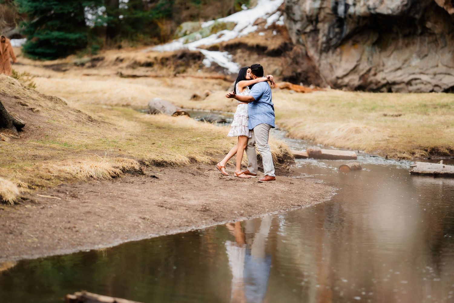 A couple embracing by a reflective pond with rocky cliffs in the background.