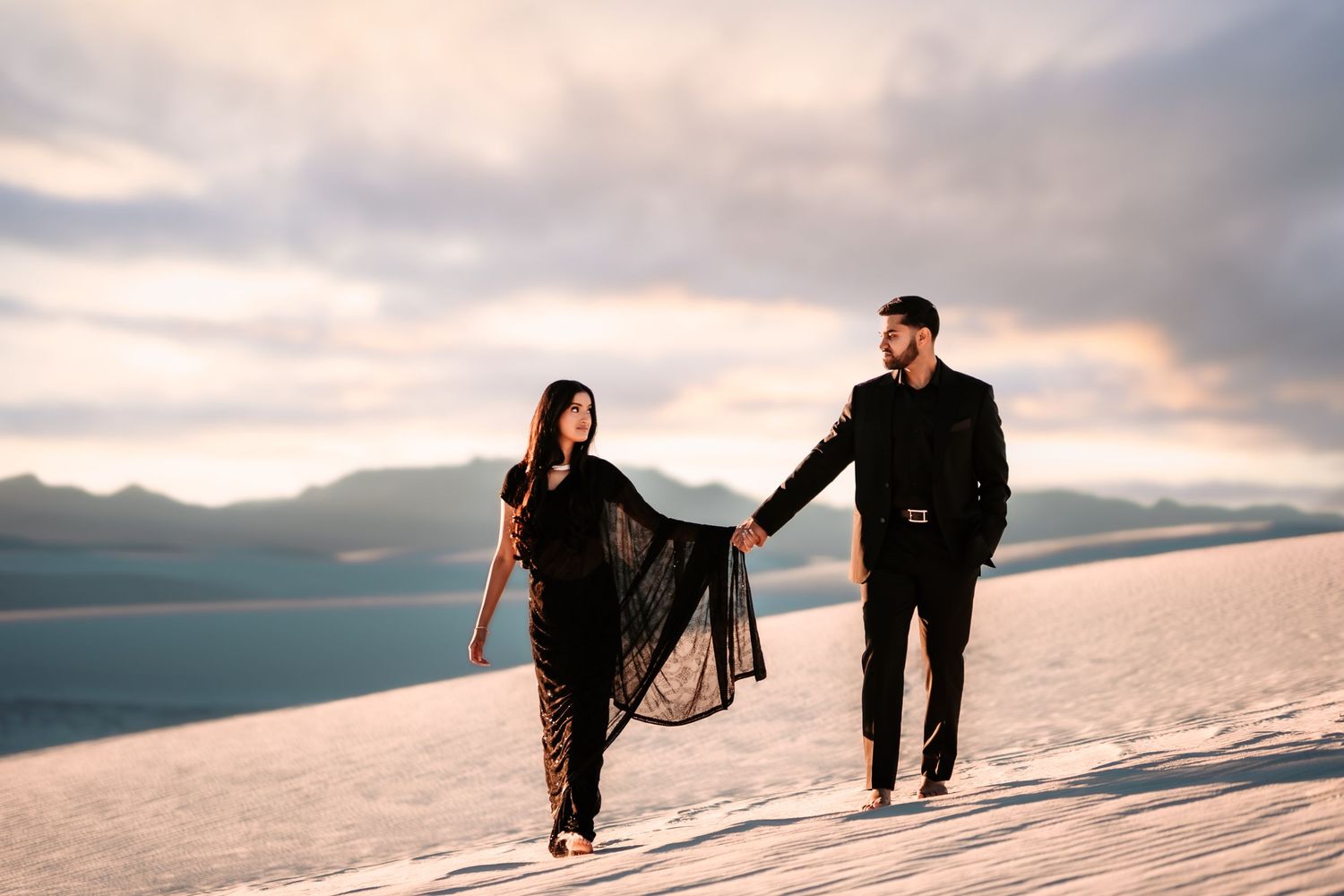 A couple walks together through a snowy mountain landscape at sunset with dramatic cloudy skies overhead.