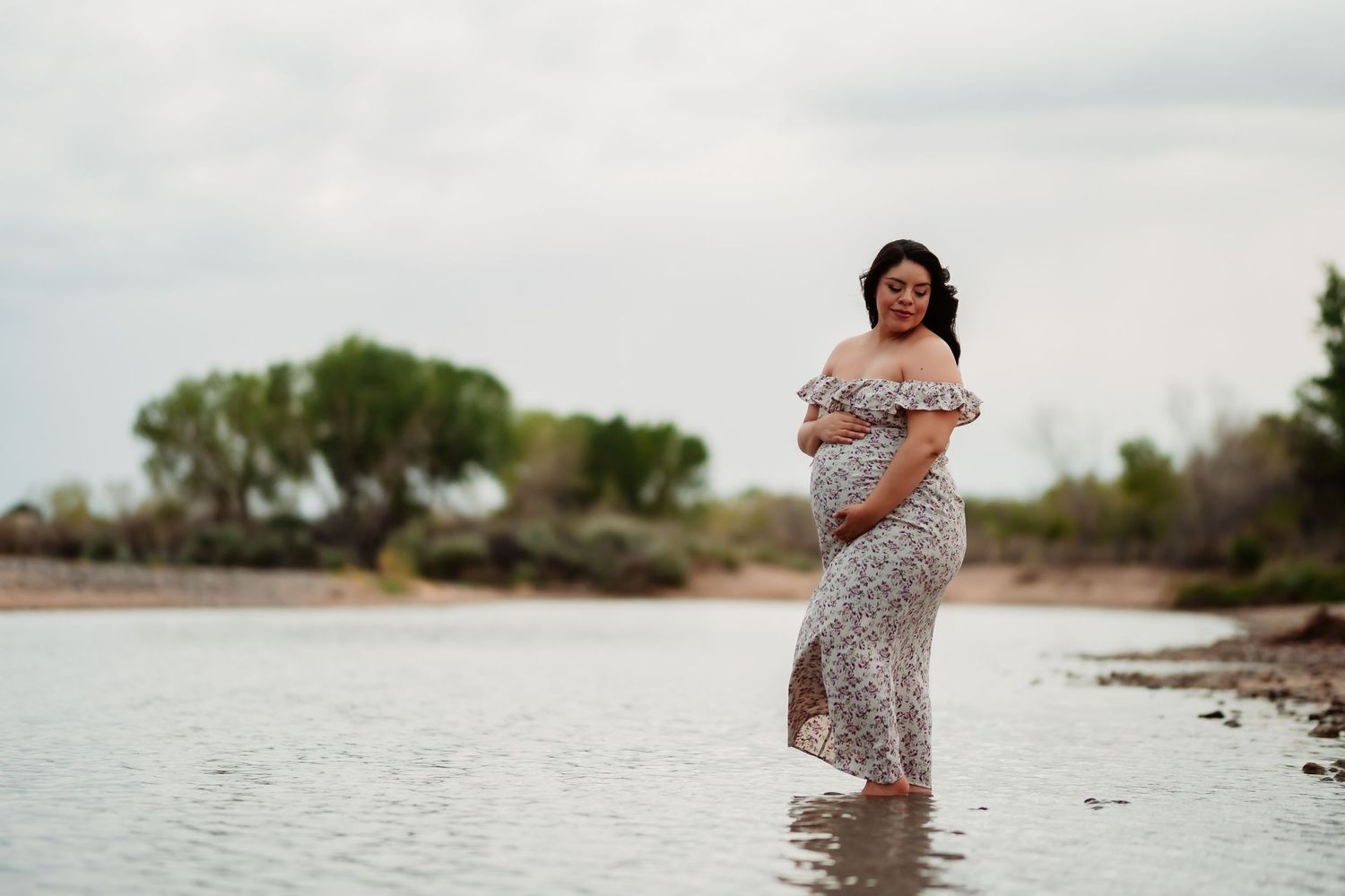 A person in a floral dress twirls joyfully in shallow water at the edge of a lake during golden hour.