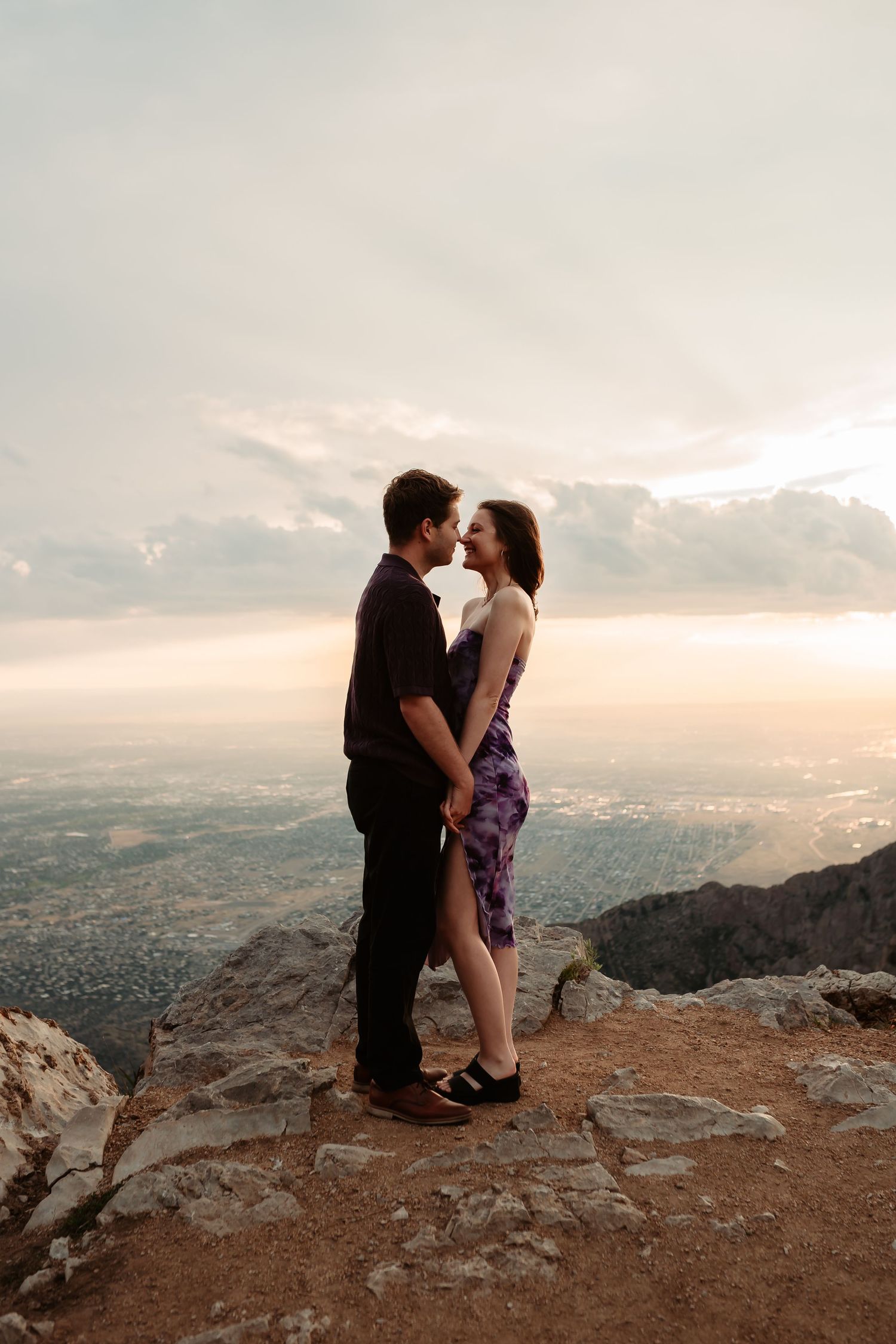 A romantic couple embraces while standing on a rocky mountain overlook during sunset.