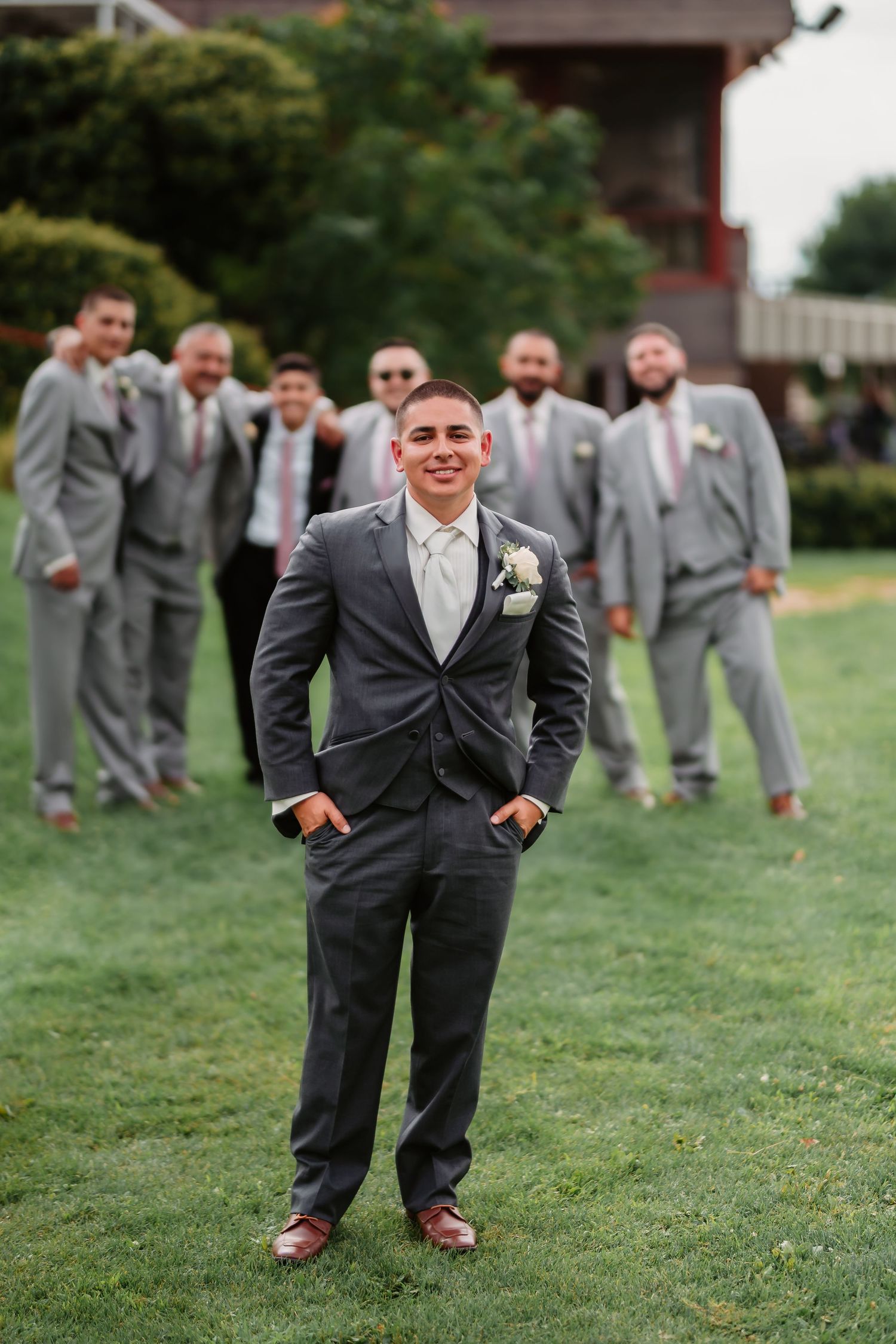 Wedding party in matching gray suits pose playfully for group photos on a grassy lawn.
