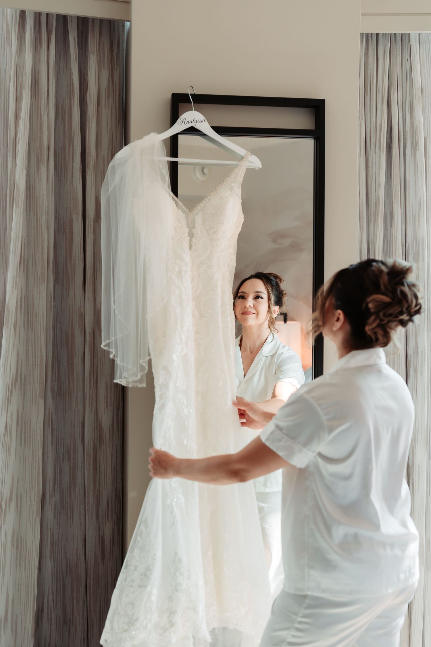 Someone in a white robe reaches to adjust a hanging wedding dress against a curtained window.