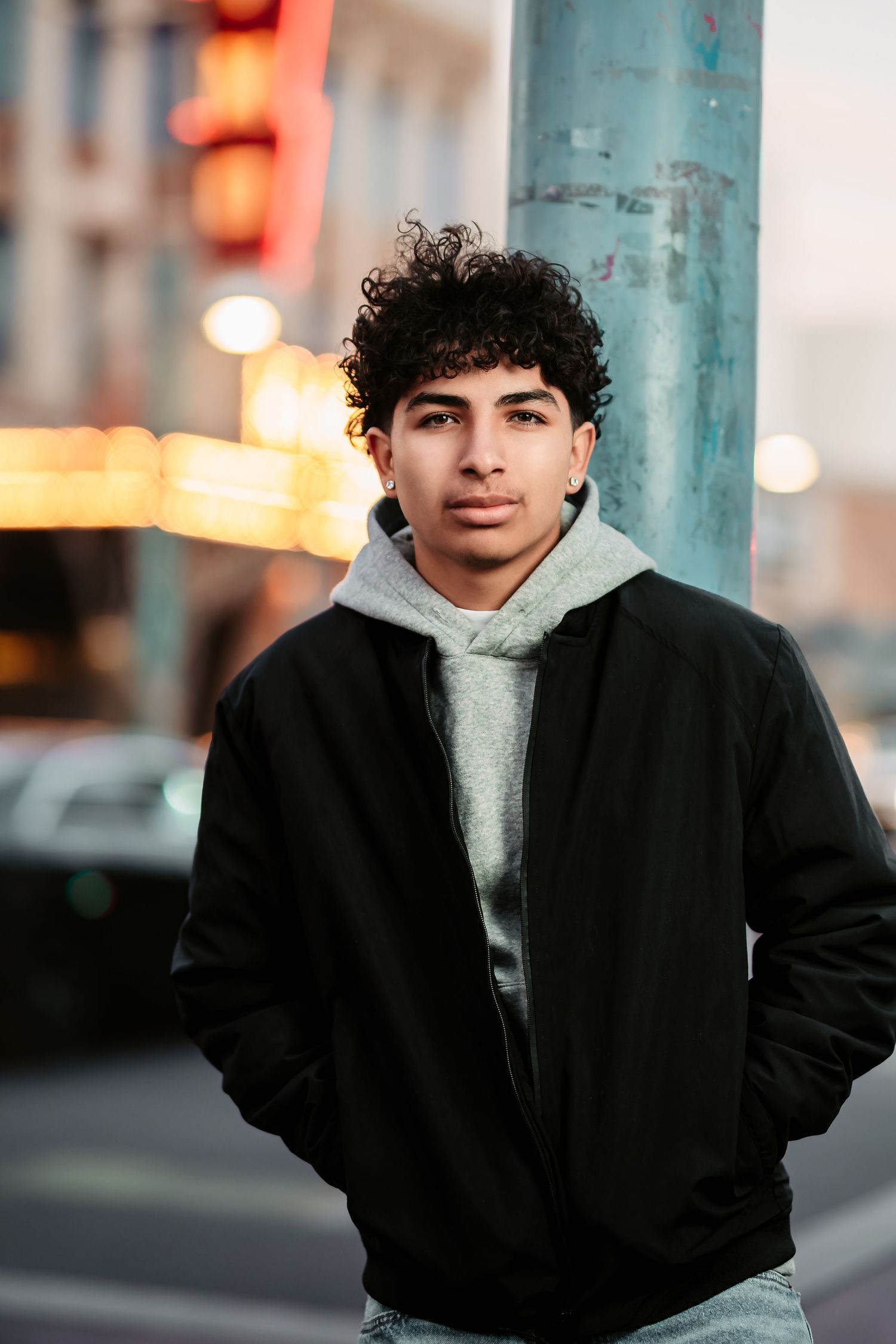 A person wearing a black jacket and white hoodie poses on a city street with blurred lights in the background.