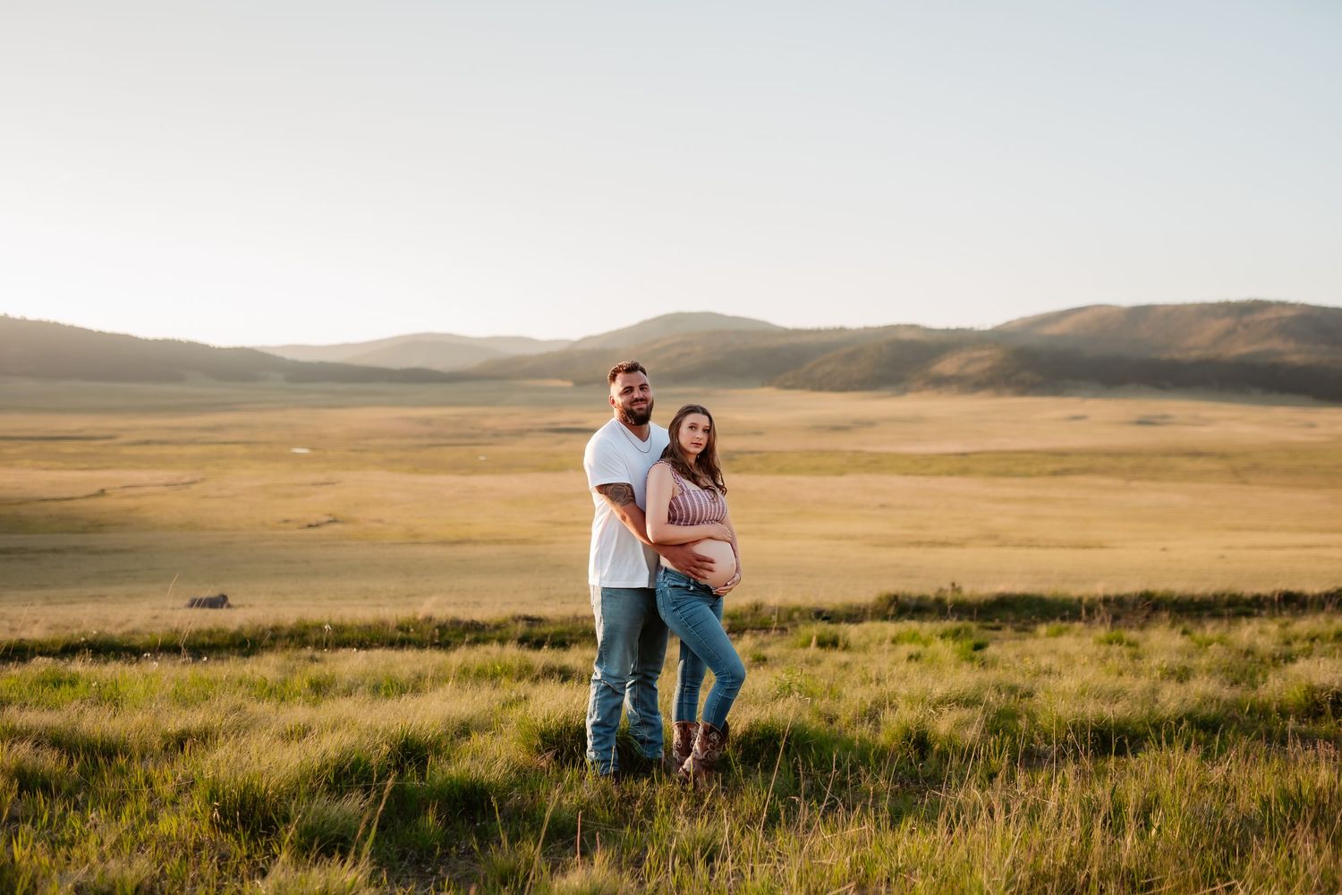 A couple walks together across a golden prairie field at sunset with mountain ranges visible in the distance.