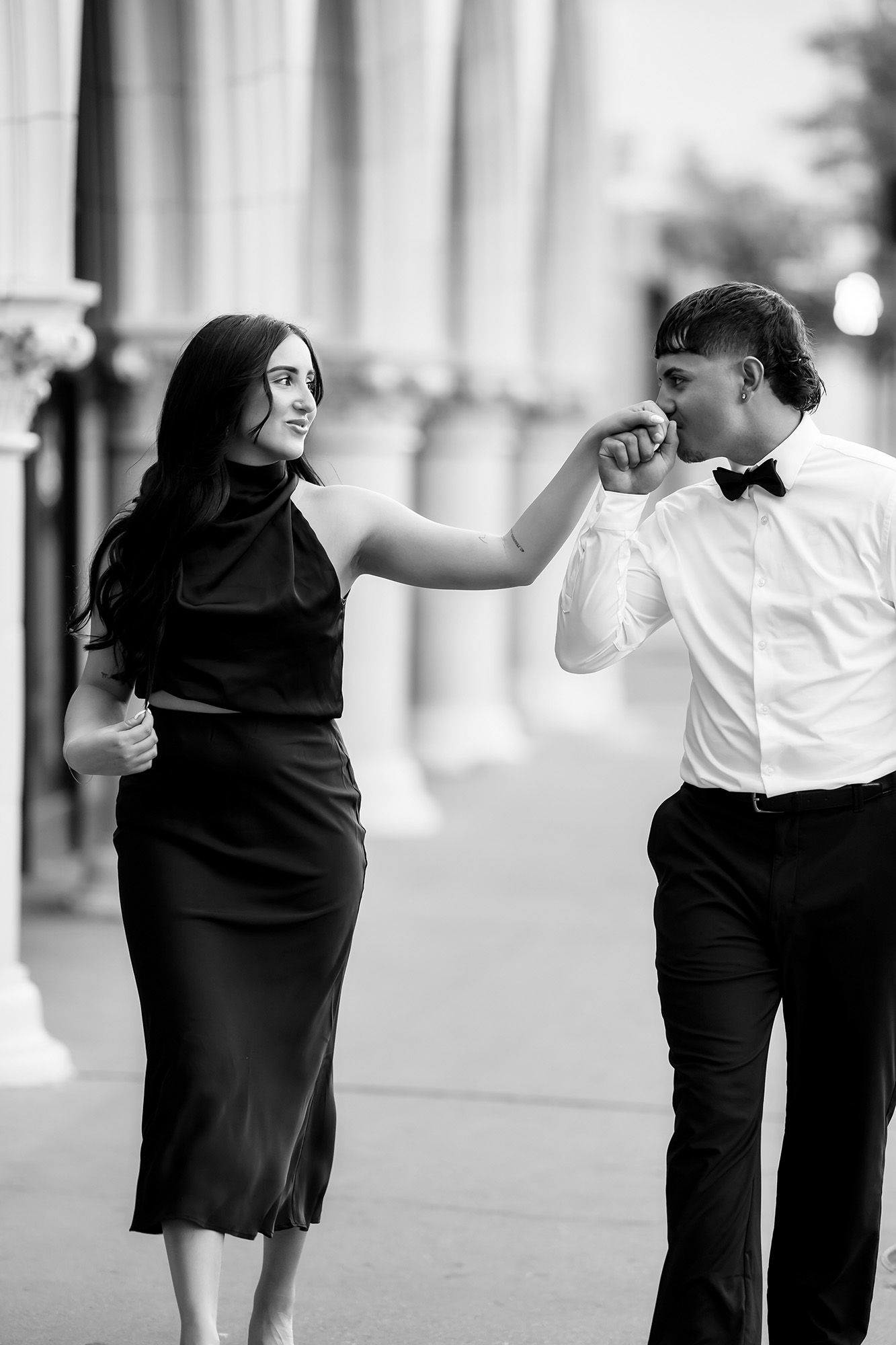 Black and white photo of elegant couple, man in bow tie kissing womans hand, wearing formal attire outdoors.