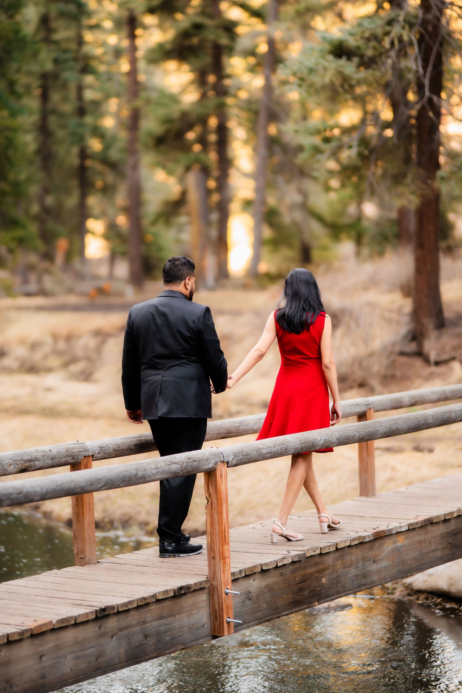 A couple walks on a wooden bridge in the forest, with her in a red dress and him in a black suit.