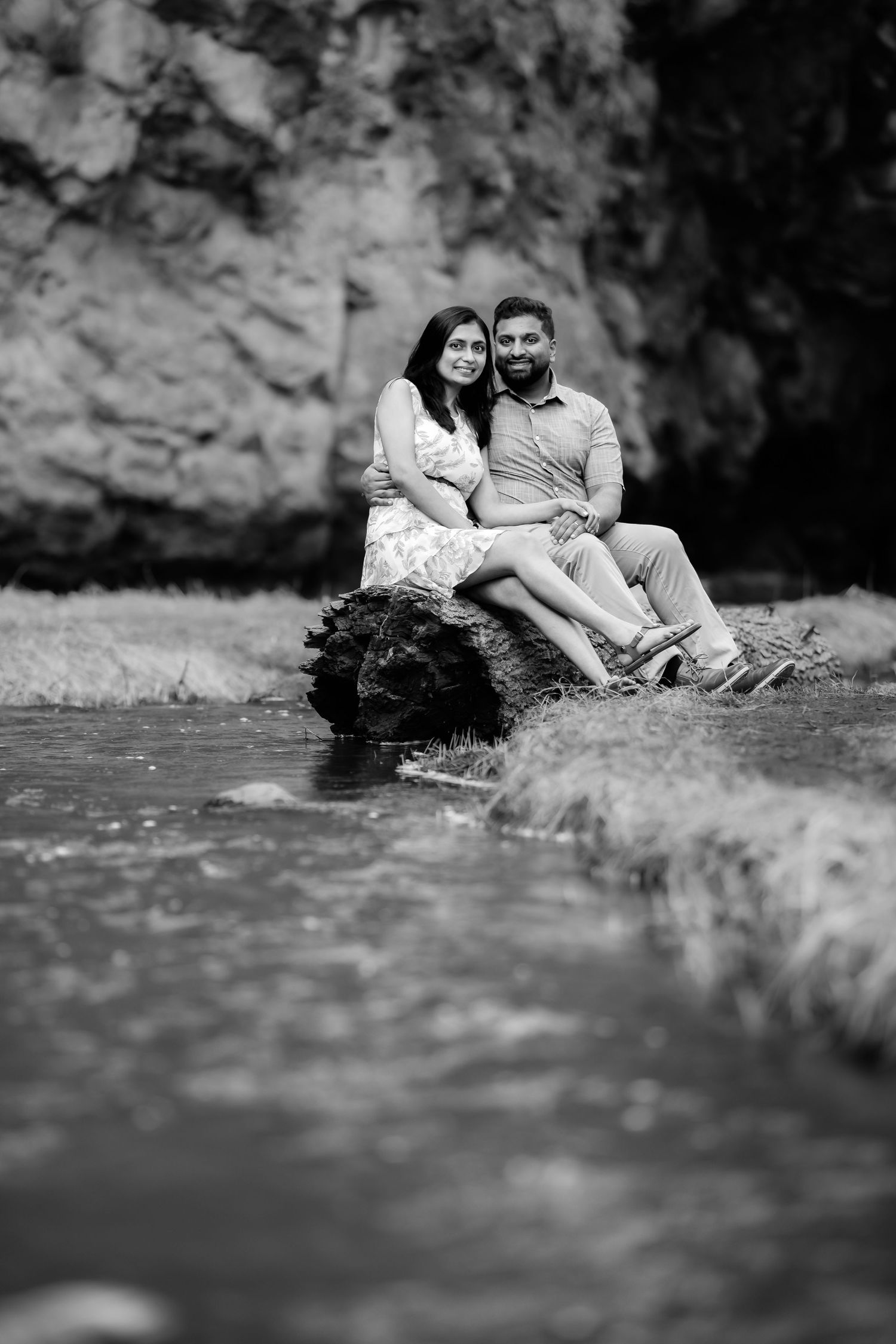 A black and white photo of a couple sitting by a stream with rocky cliffs in the background.
