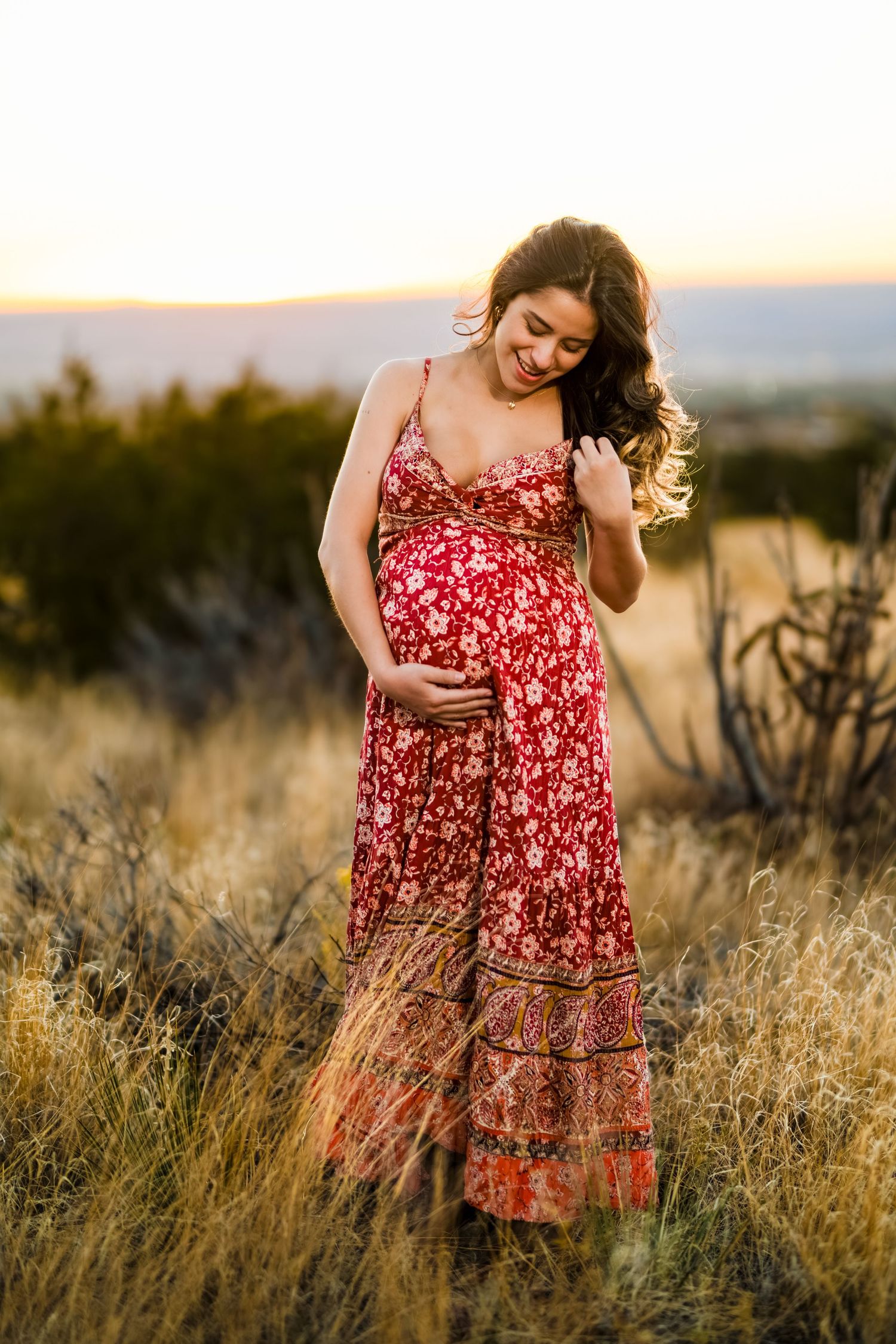 A person in a red floral dress poses gracefully in a golden field at sunset, creating a romantic outdoor portrait.