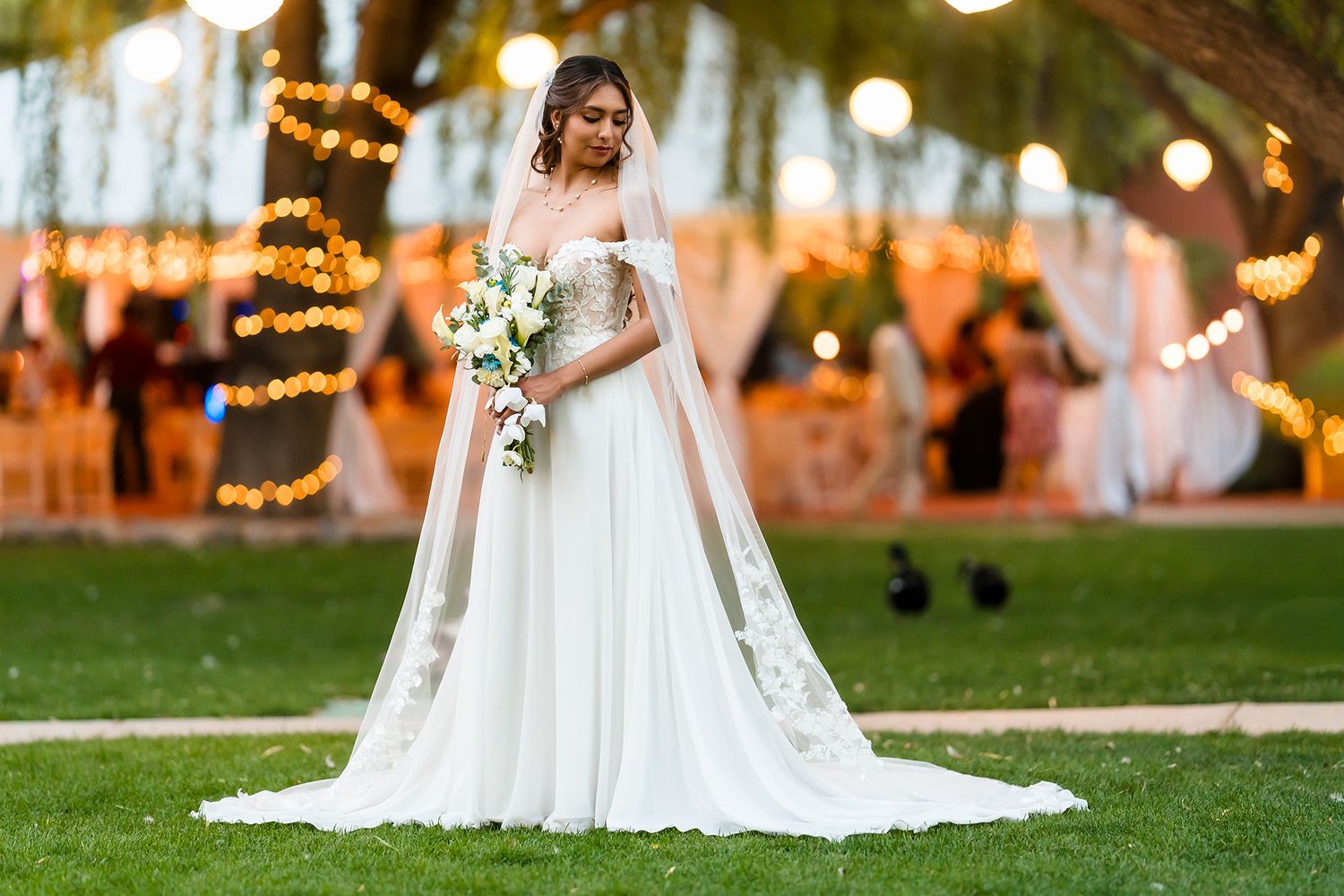 Bride in white gown holding bouquet stands on green lawn with warm fairy lights and wedding venue in background.