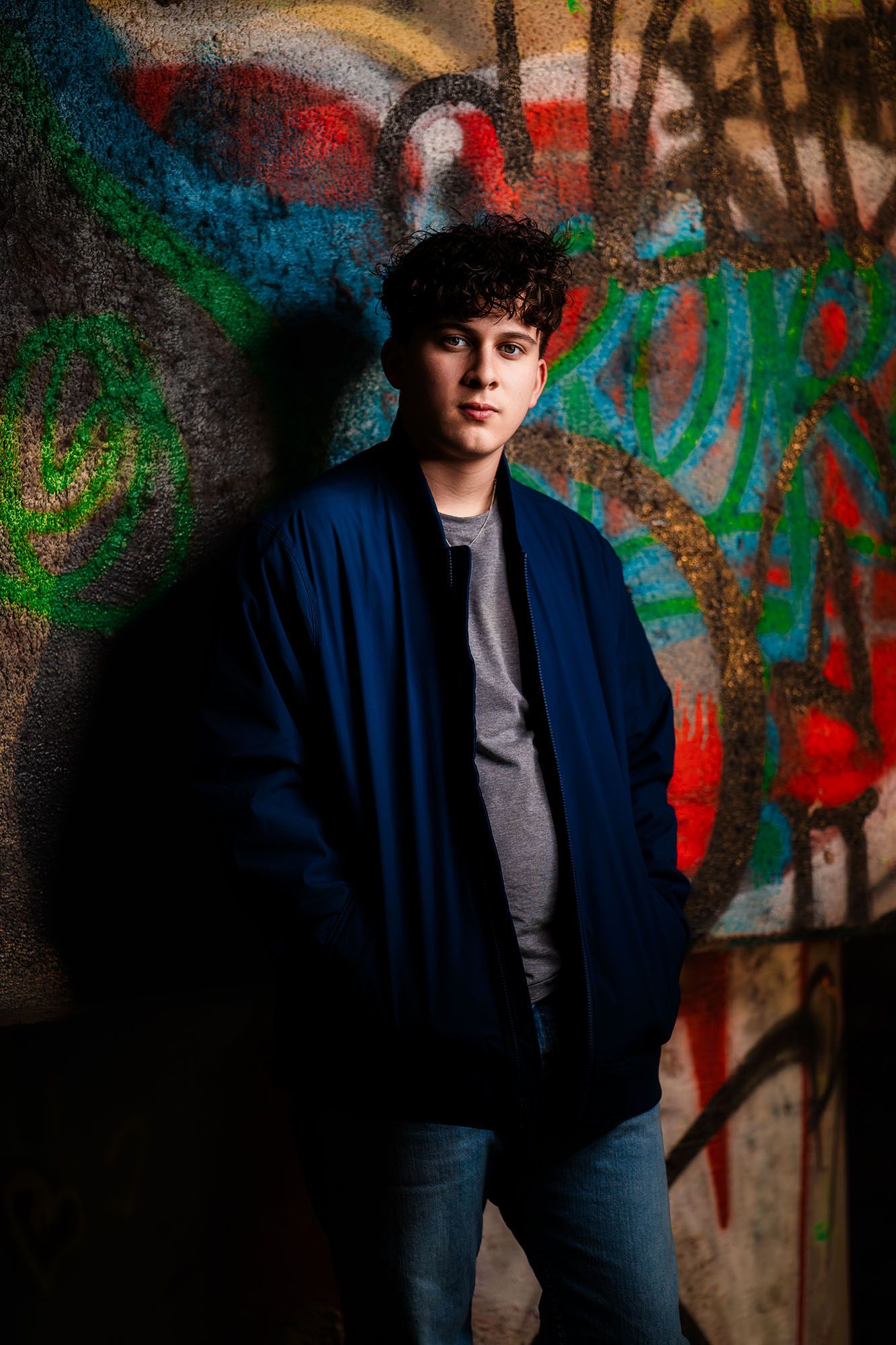 Teen boy in blue jacket leaning against colorful graffiti wall in urban street portrait.
