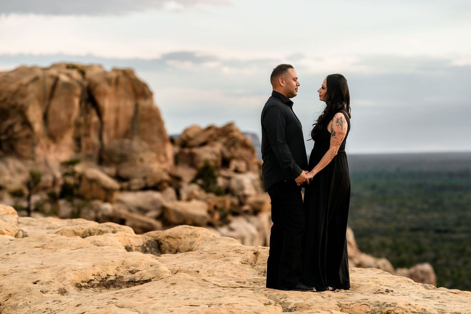 Couple in black attire holding hands on rocky desert landscape with Joshua Tree boulders and vast valley views at dusk.