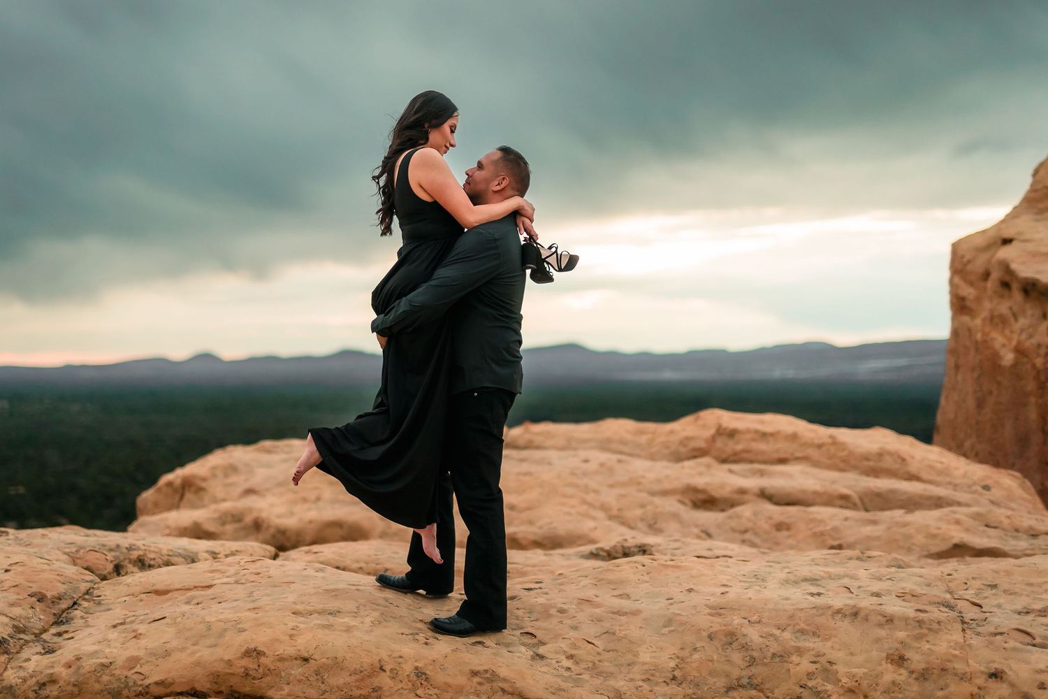 Man lifting woman in black dress on rocky cliff with dramatic stormy sky backdrop in Sedona, Arizona.