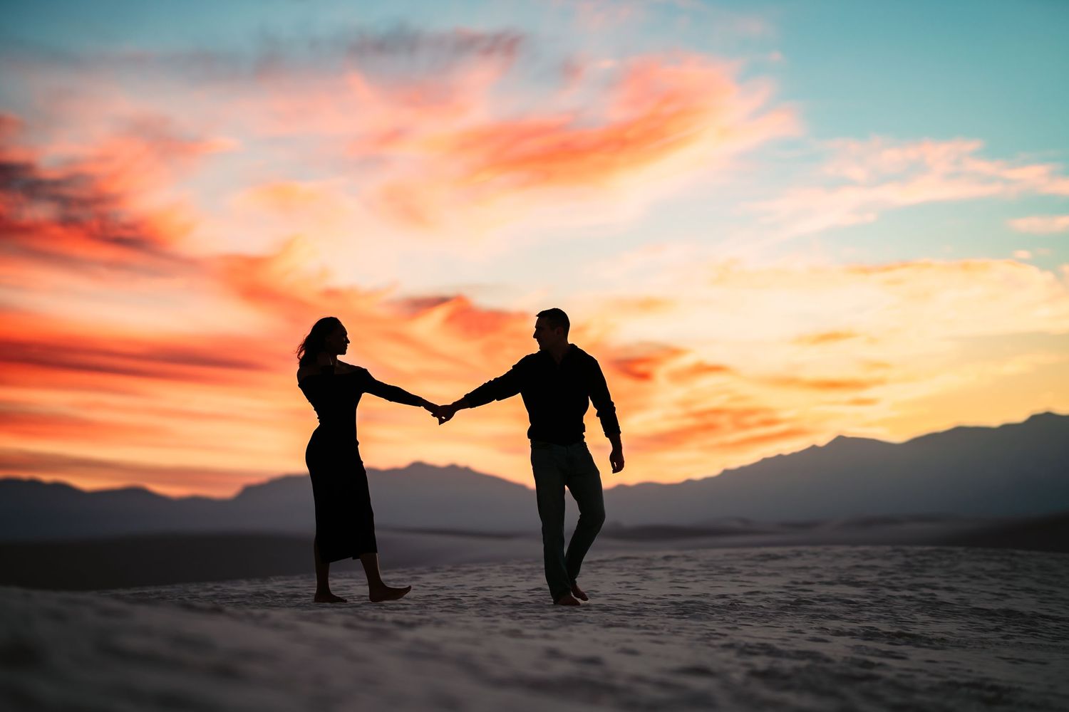 Silhouettes of people walking on sand dunes against vibrant orange and pink sunset sky with mountains in background.