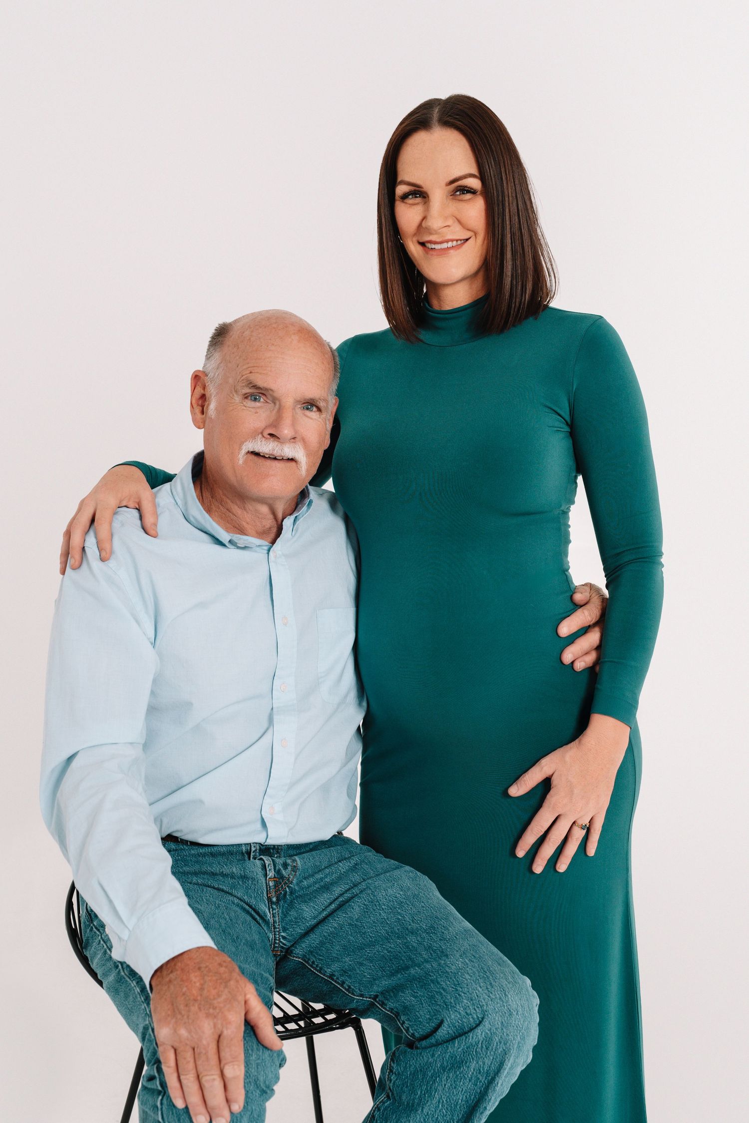 Elderly man in light blue shirt and jeans poses with woman wearing emerald green dress against white background.