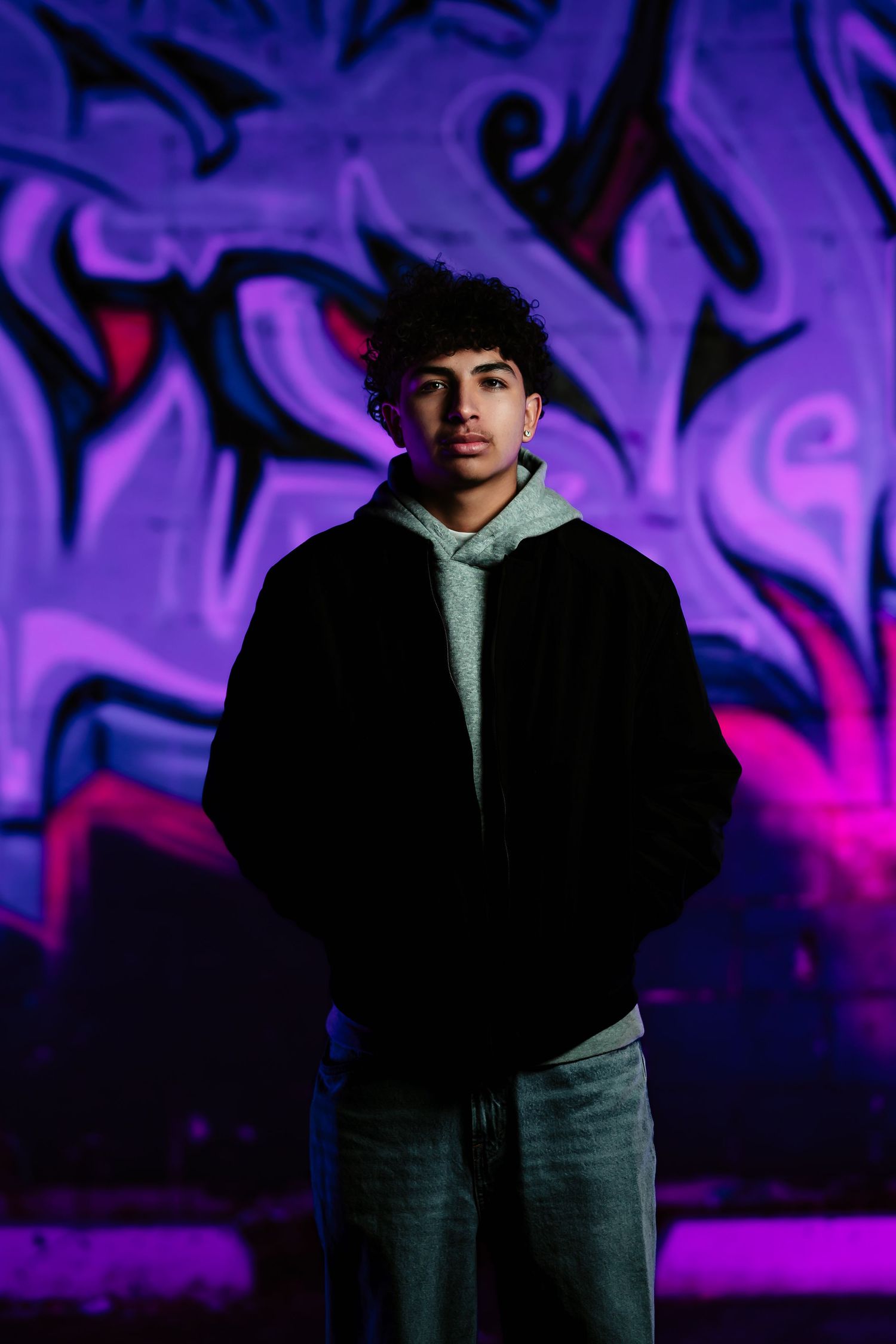 Person stands against vibrant purple graffiti wall in casual attire with dramatic lighting and urban atmosphere.