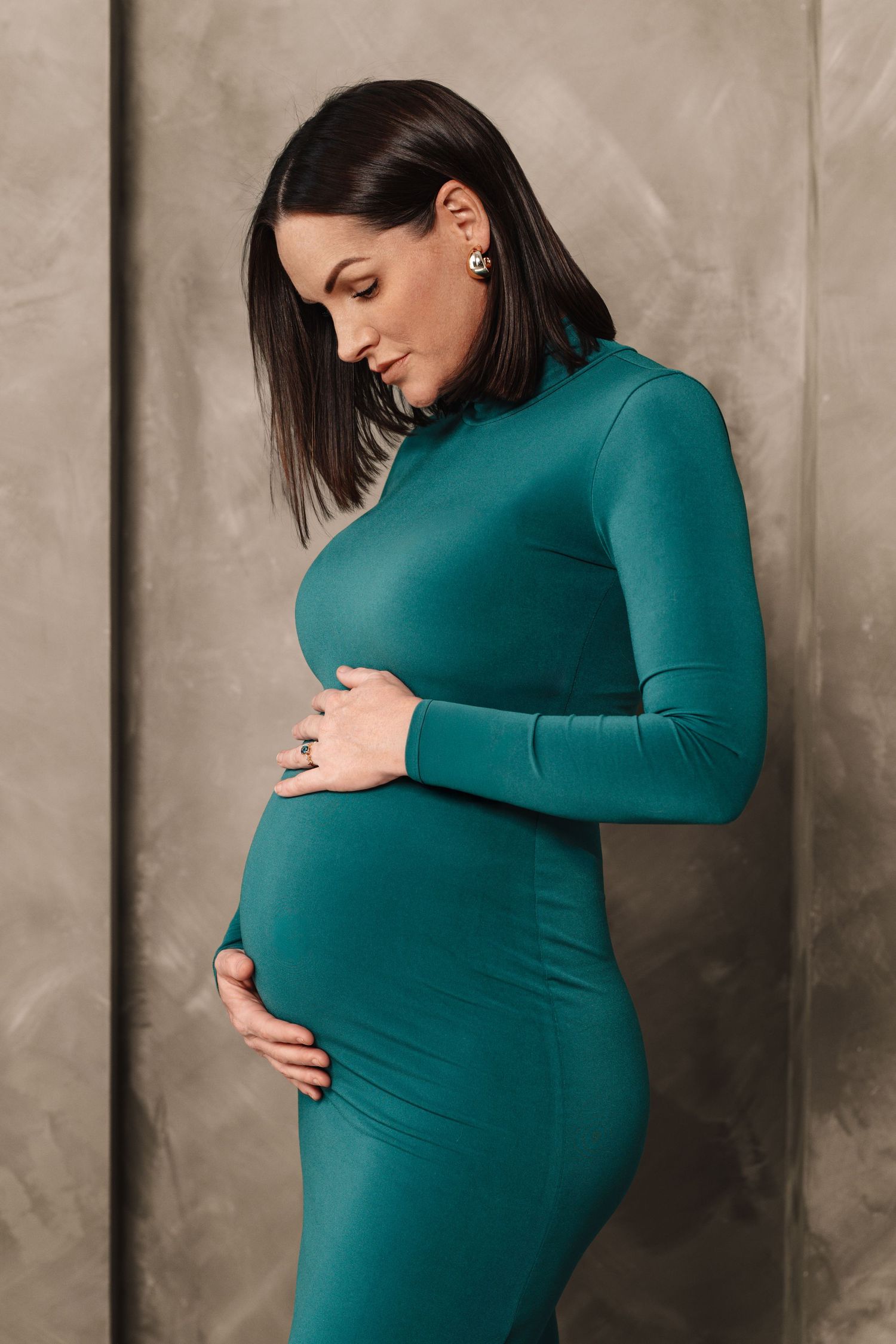 A pregnant woman in a teal turtleneck dress poses against a gray backdrop in a series of maternity photos.