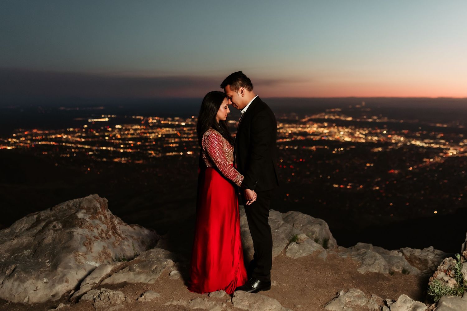 A romantic couple enjoys sunset views on a hilltop overlooking a city's glittering lights in the distance.