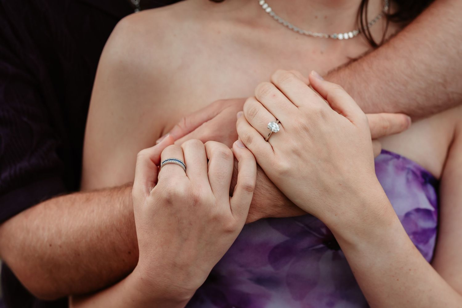 Close-up of hands wearing wedding rings with purple dress fabric and pearl necklace details.