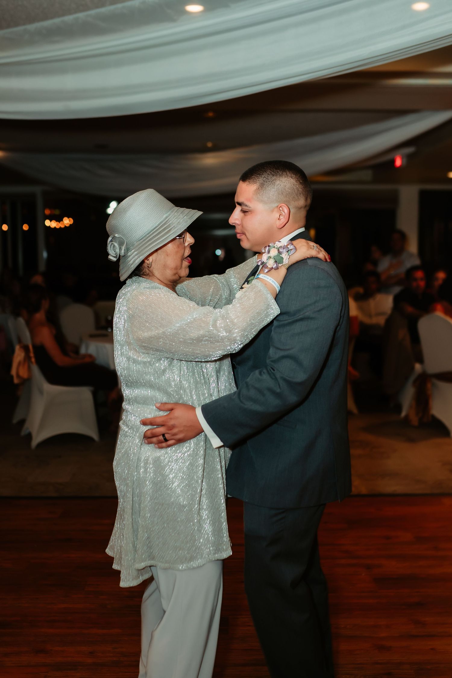 A couple shares a tender dance in formal wear under draped ceiling decor at an indoor reception venue.
