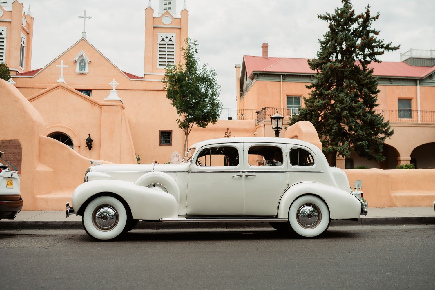 A classic white vintage car parked in front of a pink adobe church building.