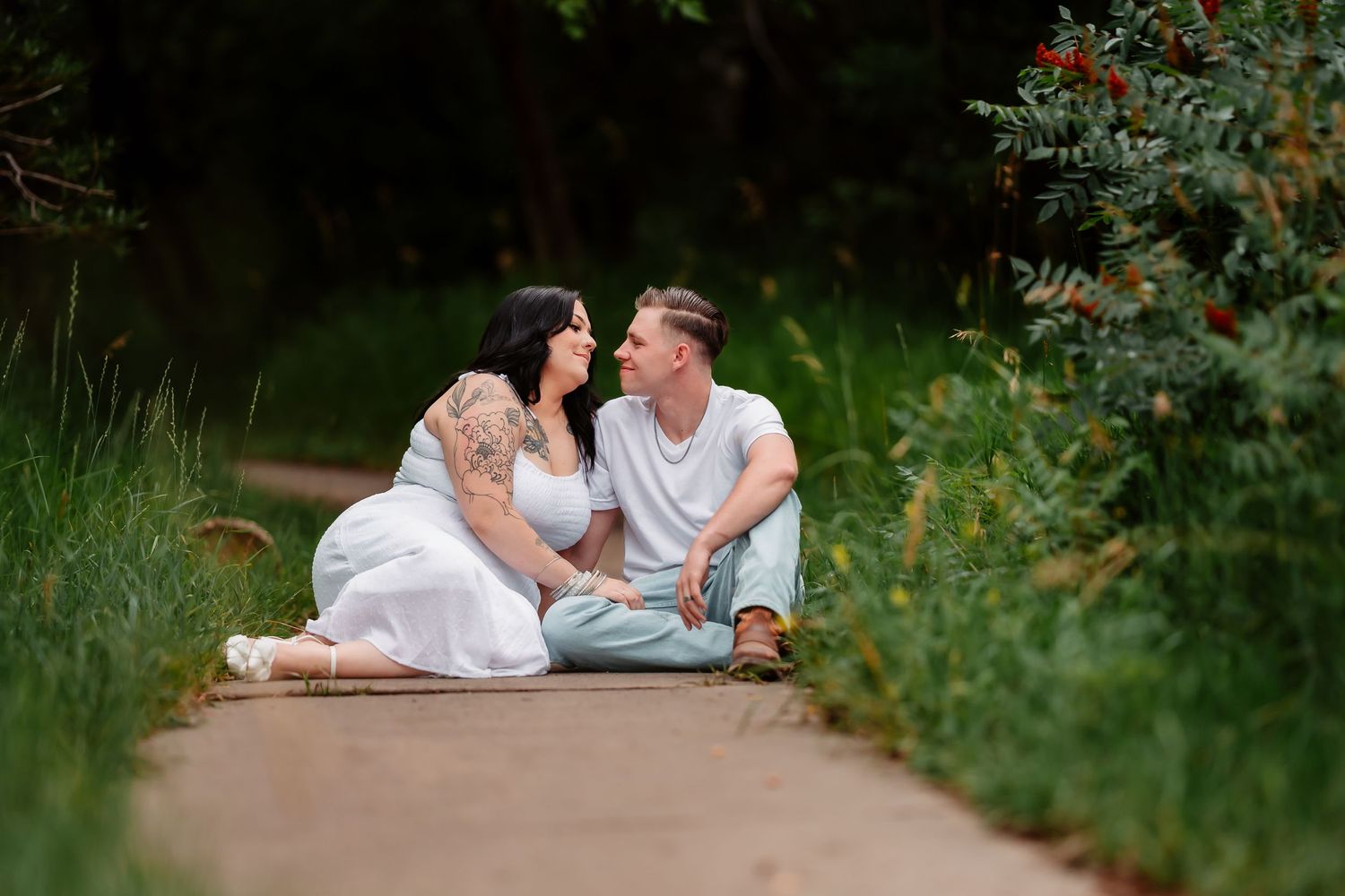 Two people in white clothing sitting together on a path surrounded by lush green foliage on a summer day.