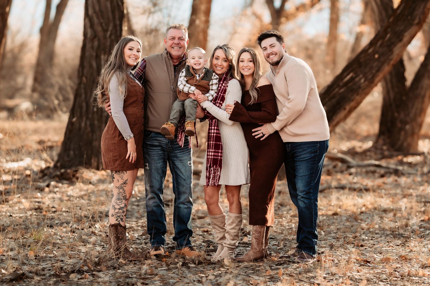 Family of six posing outdoors in fall foliage, wearing coordinated earth-tone outfits, smiling together among autumn trees.