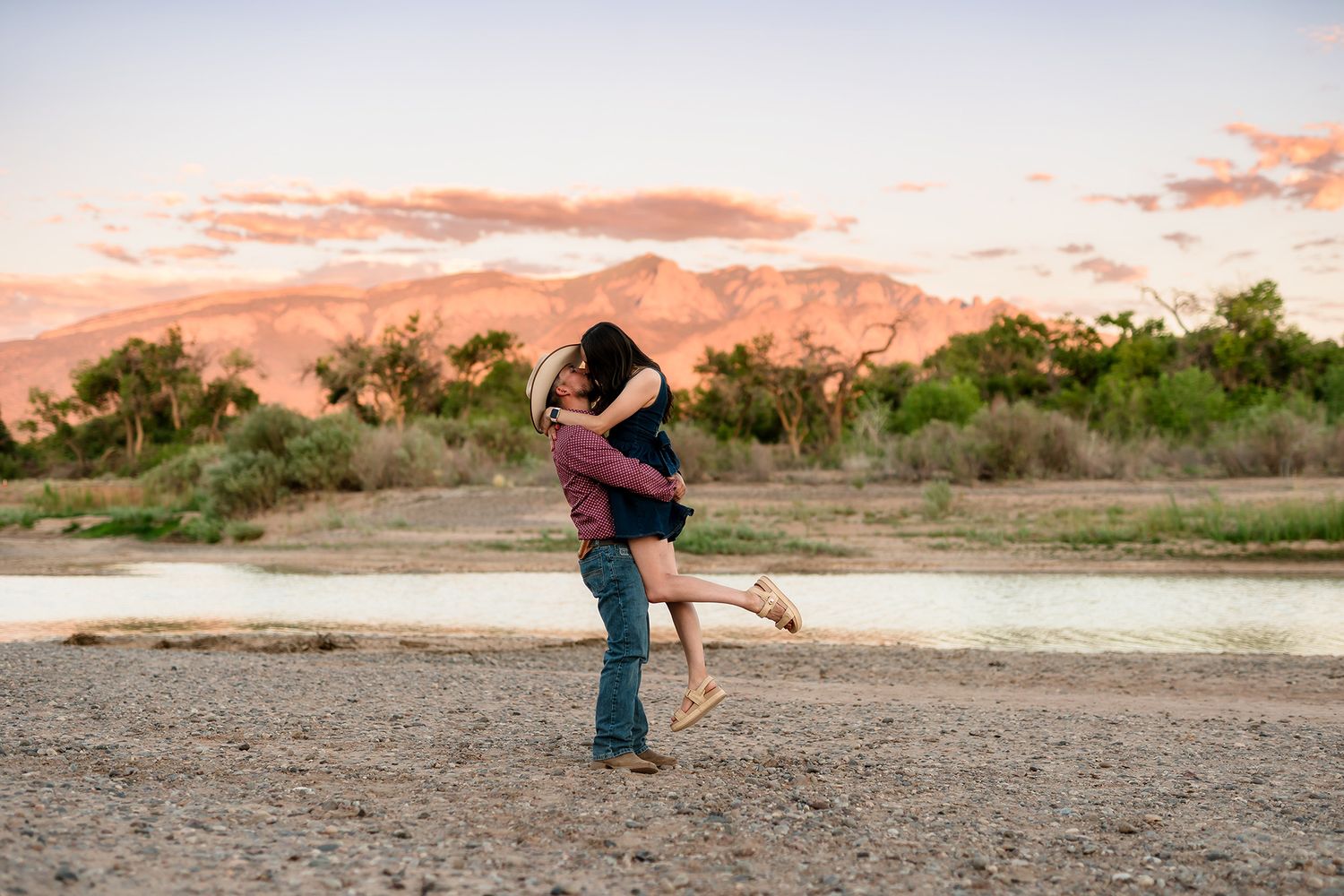 Couple sharing romantic embrace at sunset near a river with mountains and colorful sky in the background.