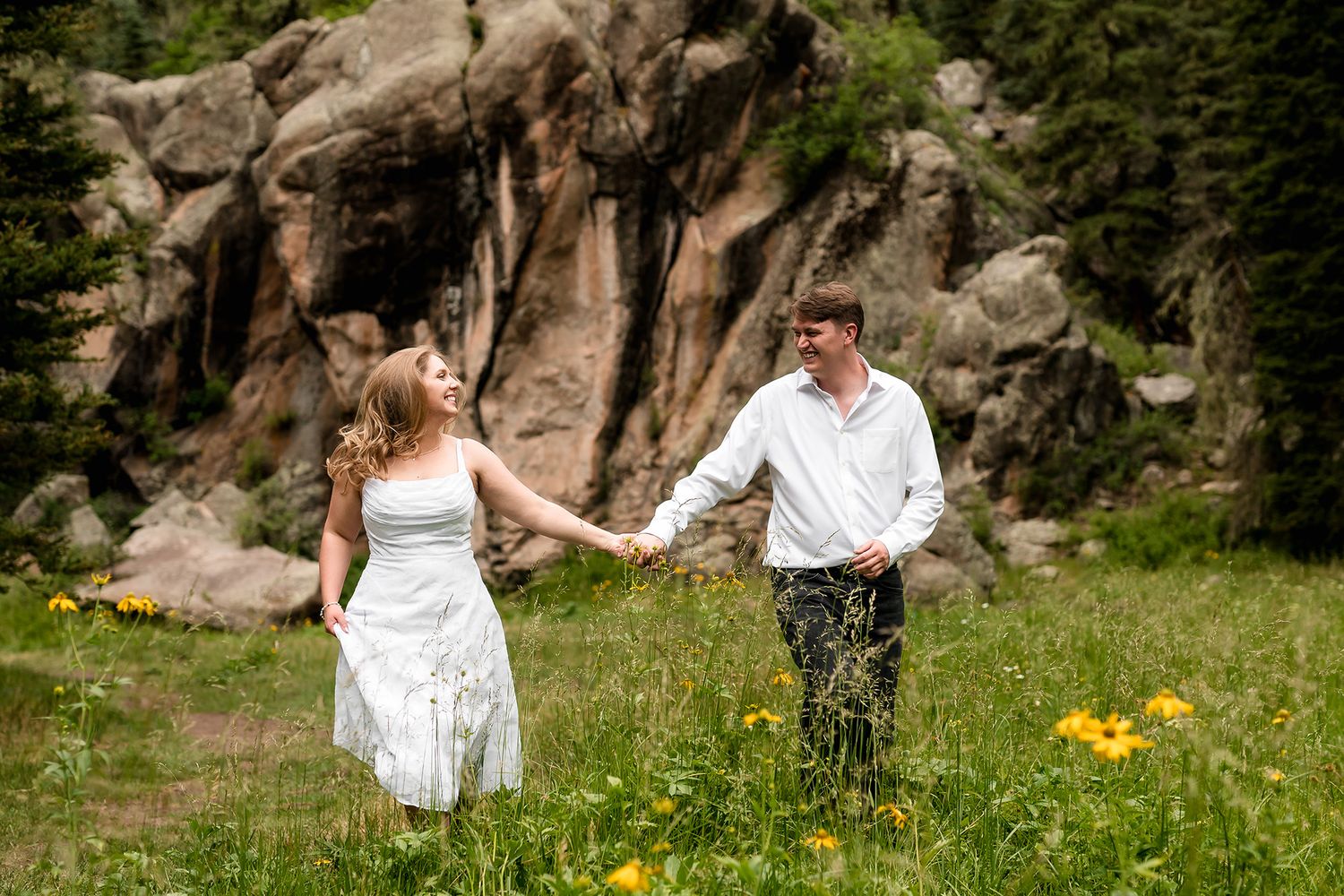 Couple holding hands walking through wildflower meadow with rocky mountain backdrop on wedding day.