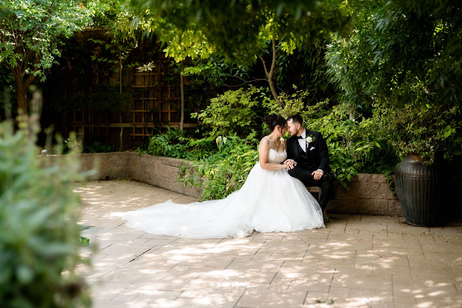 Bride and groom share a romantic moment seated in a sunlit garden courtyard surrounded by lush green trees.