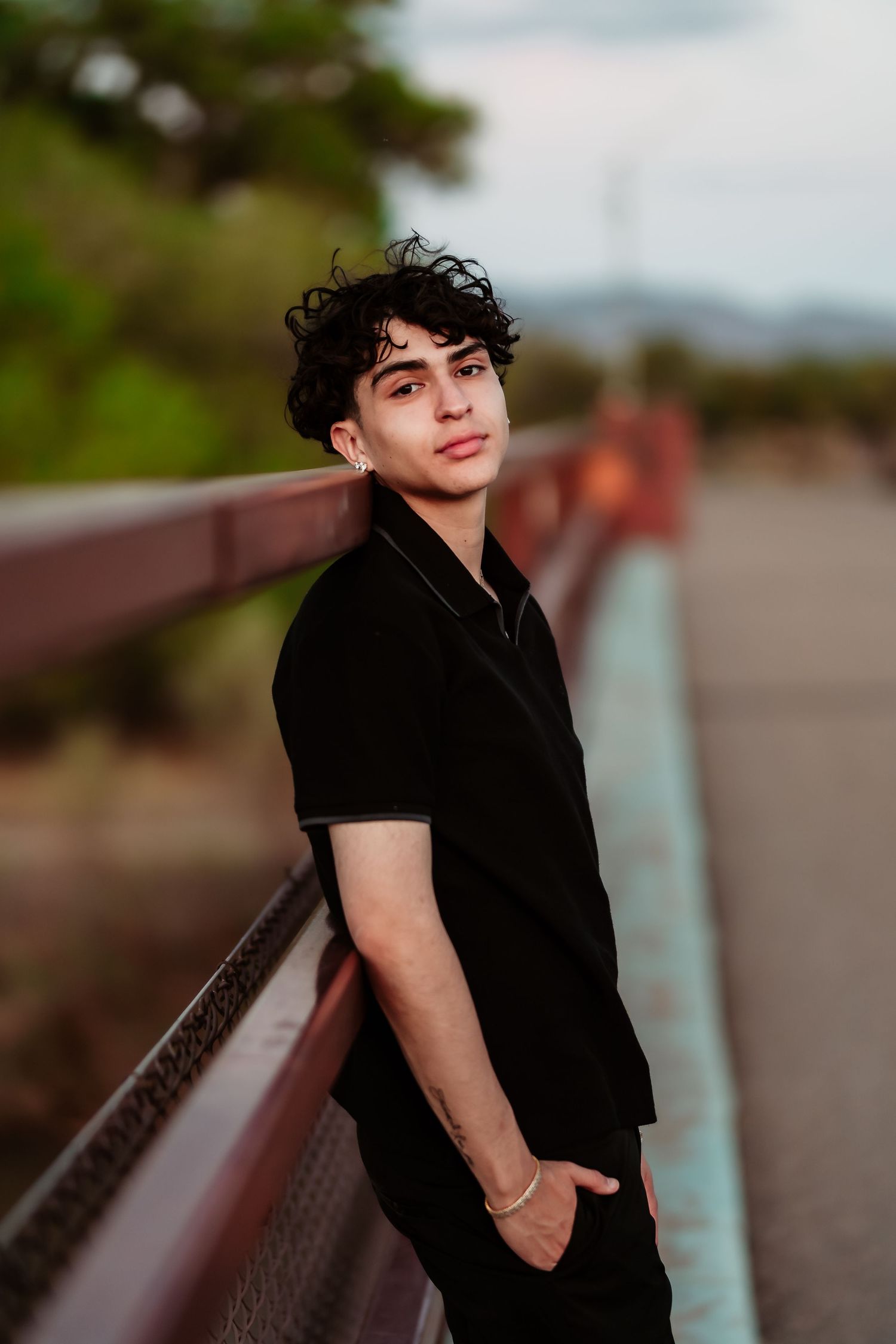 A figure in all black clothing leans against a railing during golden hour with curly dark hair and casual pose.