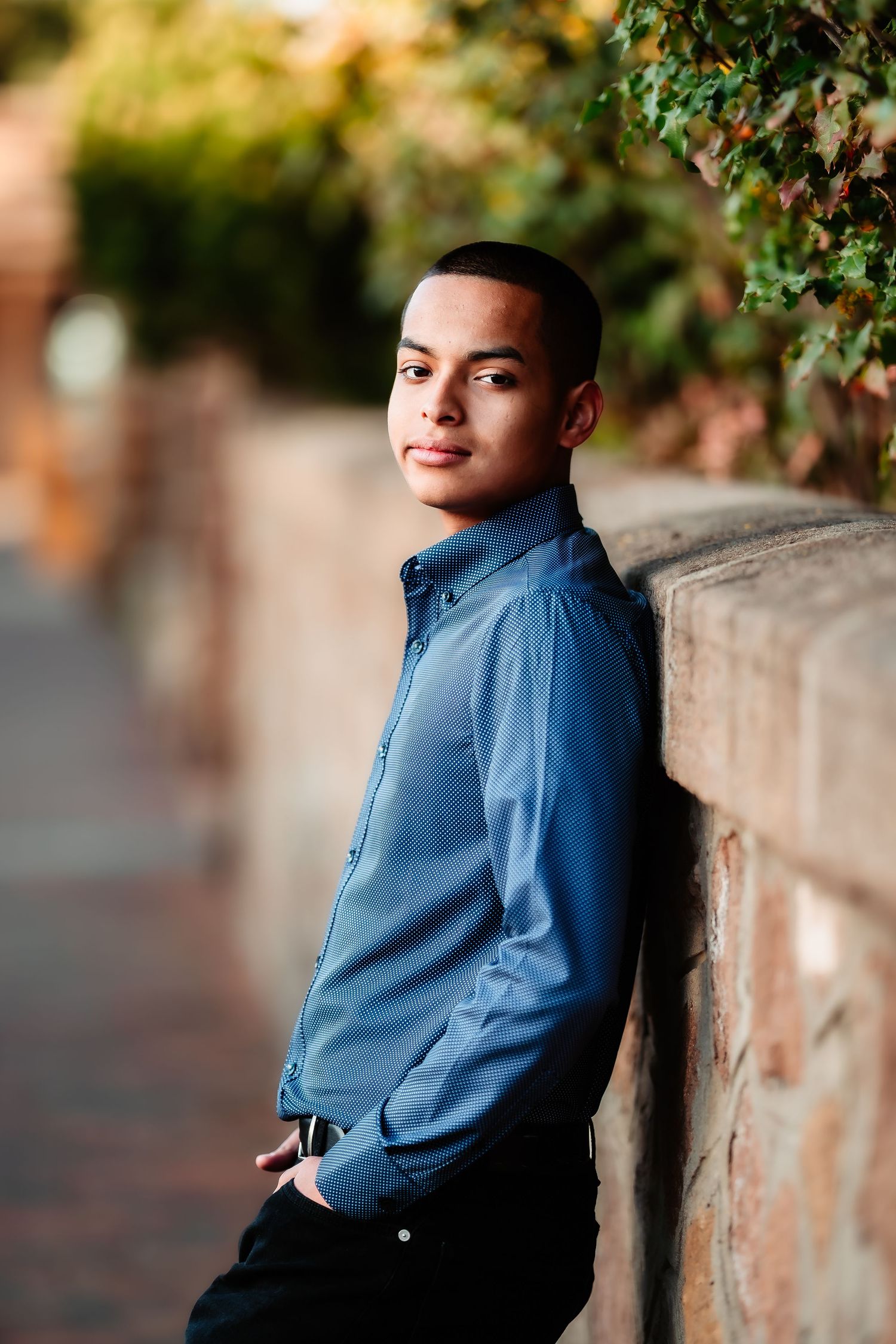 Young person in blue button-down shirt leaning against brick wall in outdoor setting with soft natural lighting.