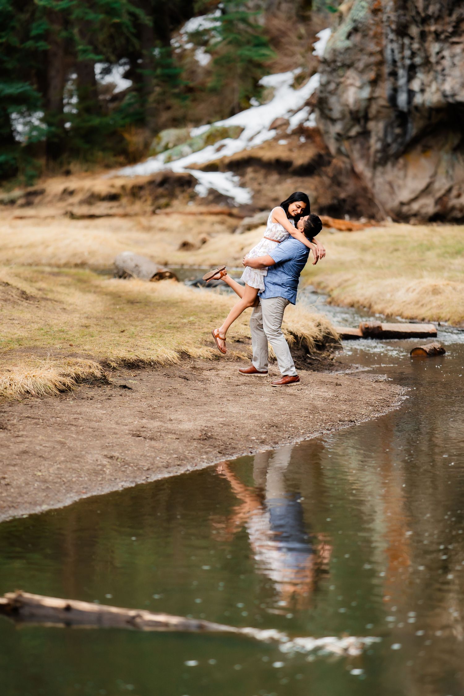 Playful couple spins together by reflecting mountain stream with snowy cliffs in background.