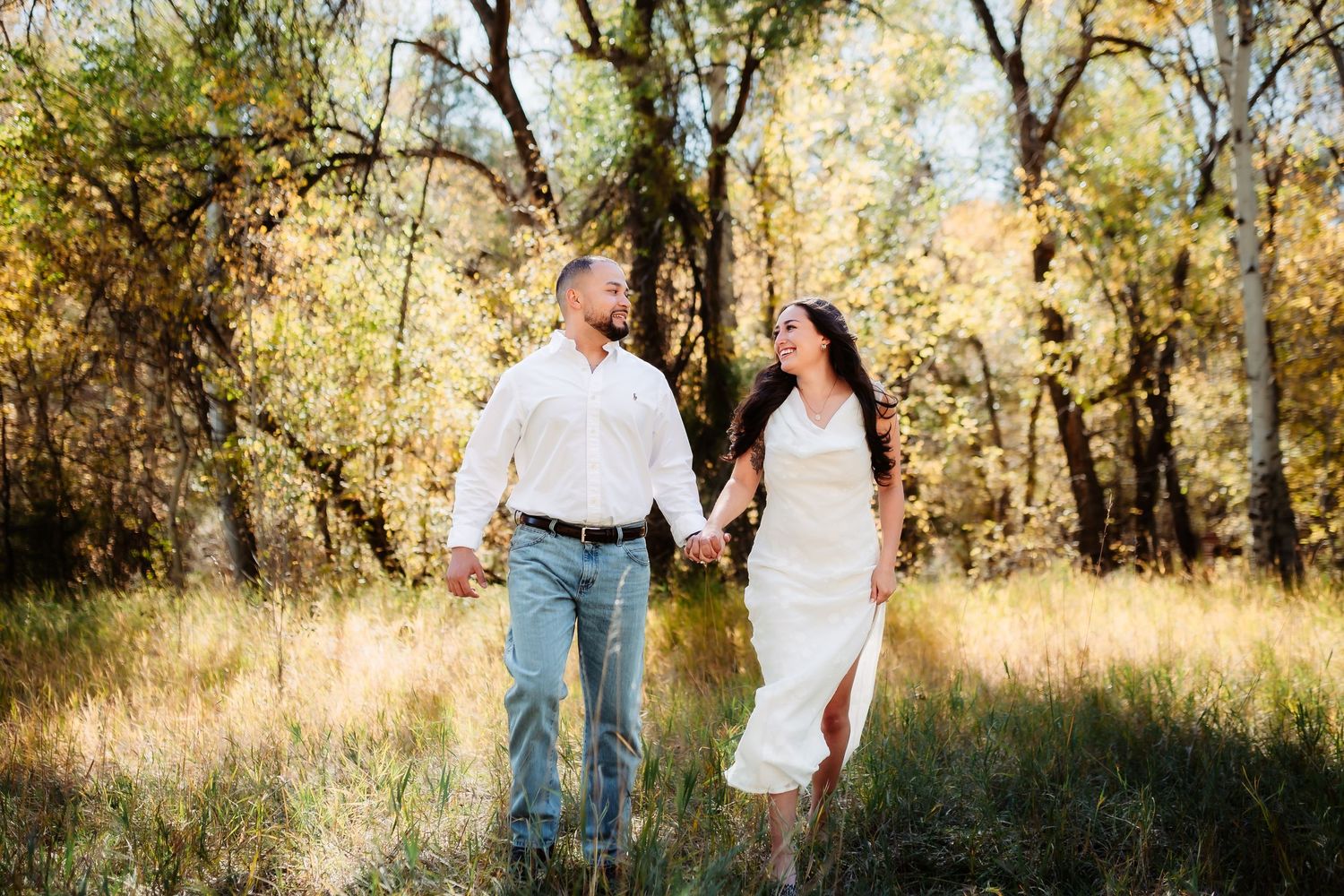 A couple walks hand in hand through a sunlit autumn forest during a romantic outdoor photo session.