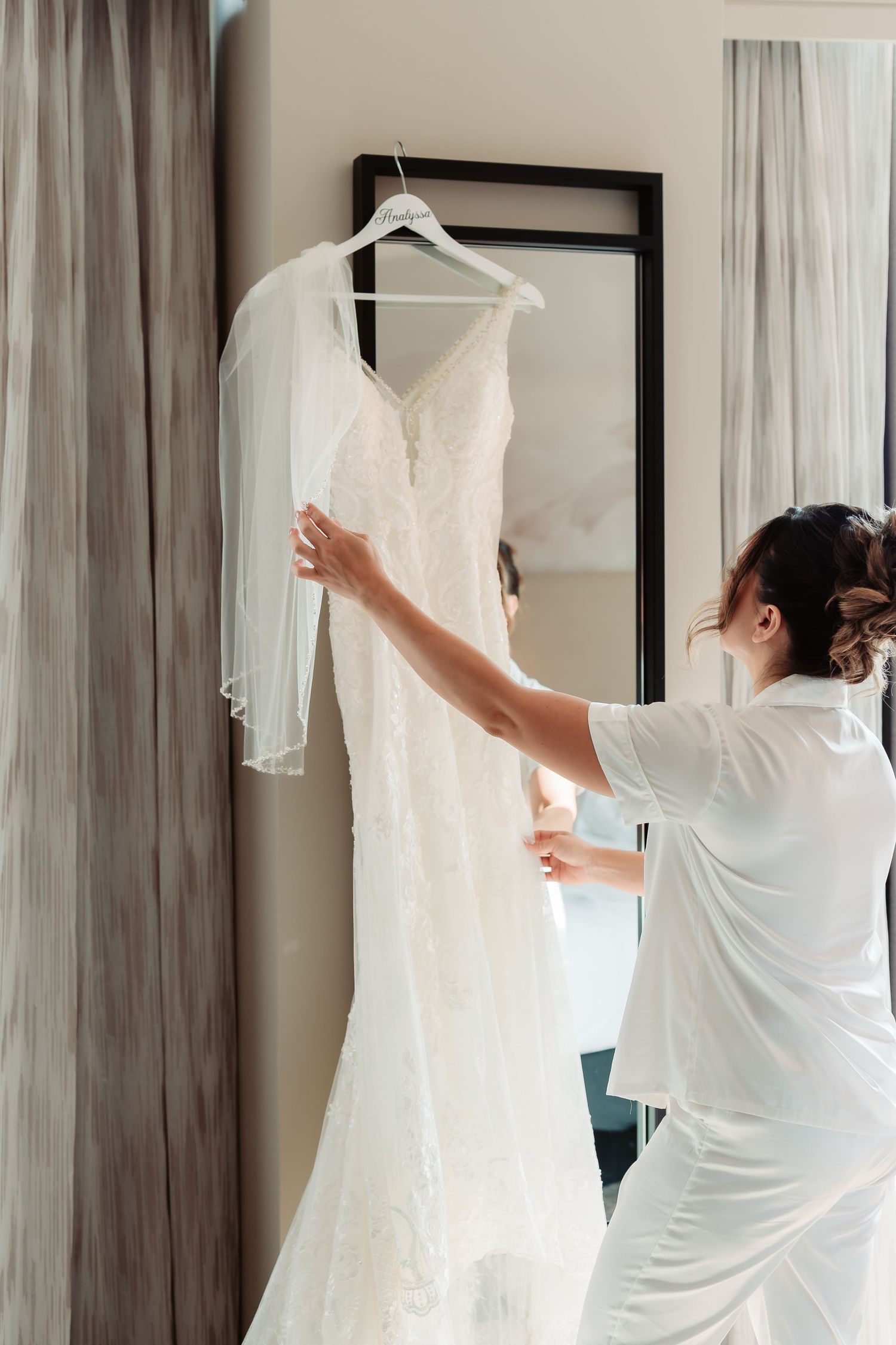 A white wedding dress hangs from a hanger in a hotel room with soft lighting and curtains.