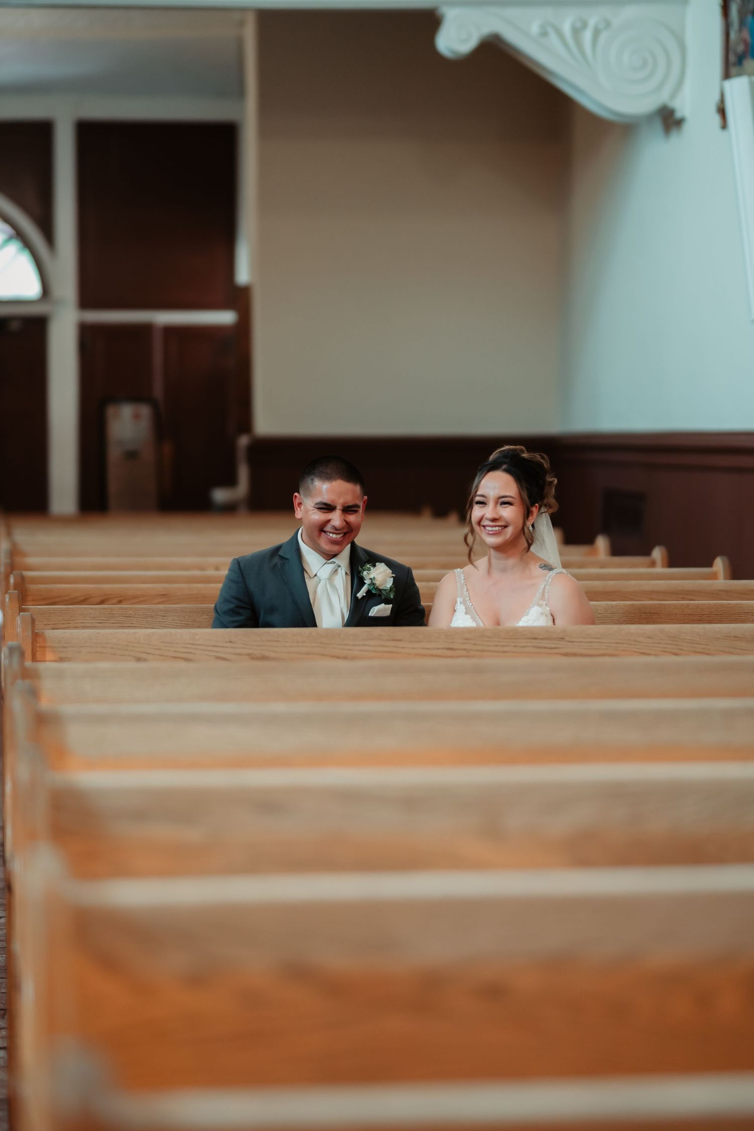 A bride and groom share a joyful moment while sitting in wooden church pews during their wedding ceremony.