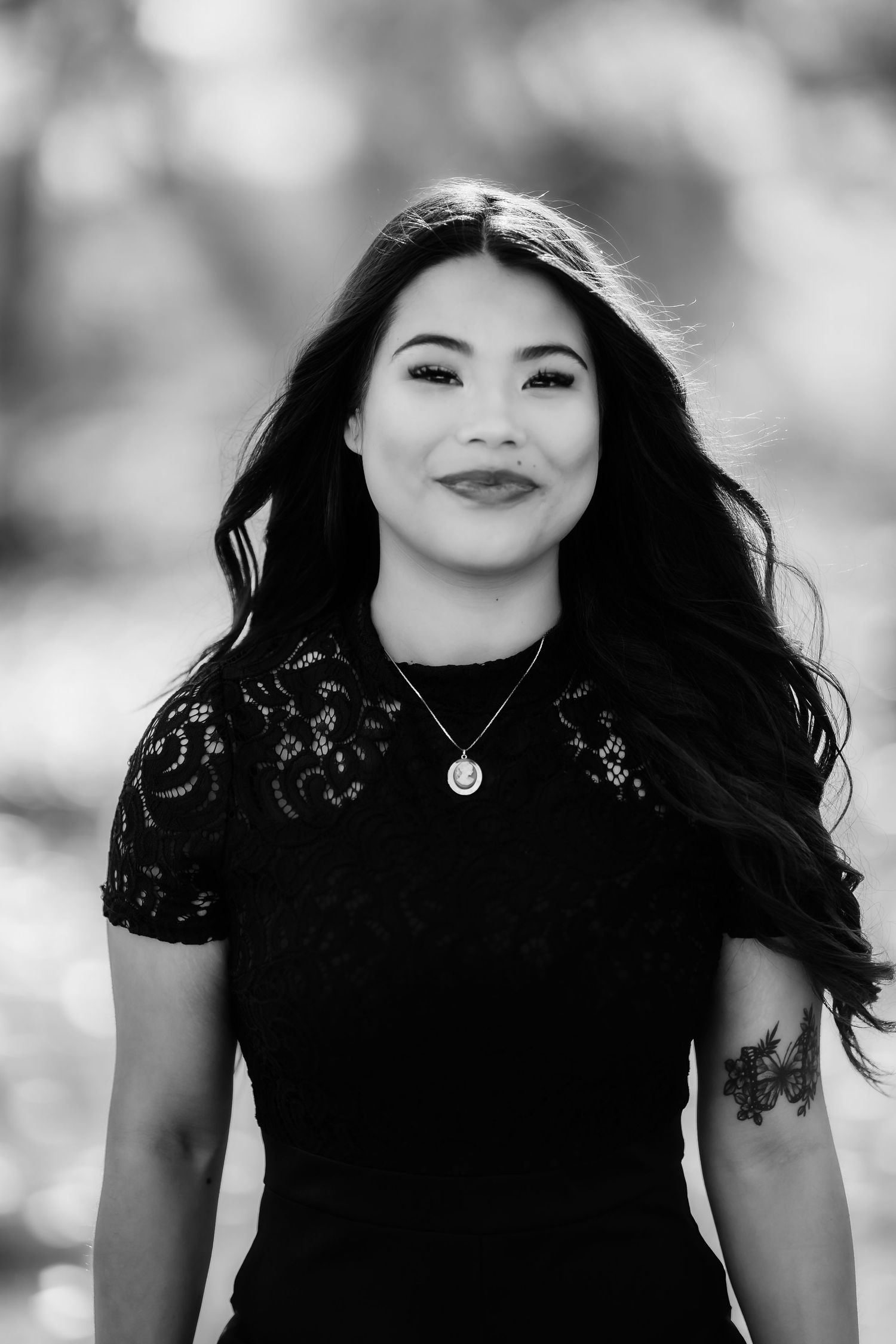 Black and white portrait of a woman with flowing dark hair wearing a black lace top in a dreamy outdoor setting.