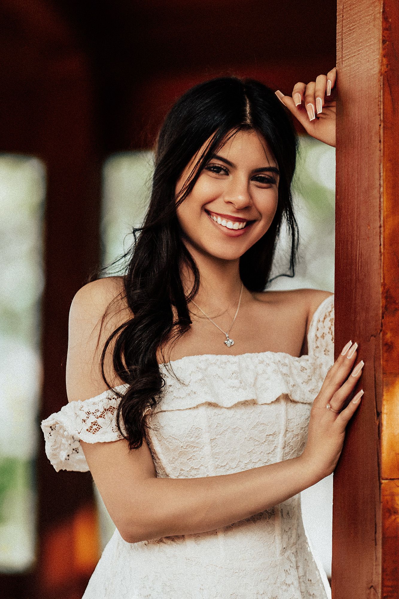 A woman in a white lace off-shoulder dress poses against a wooden pillar in a warm-toned setting.