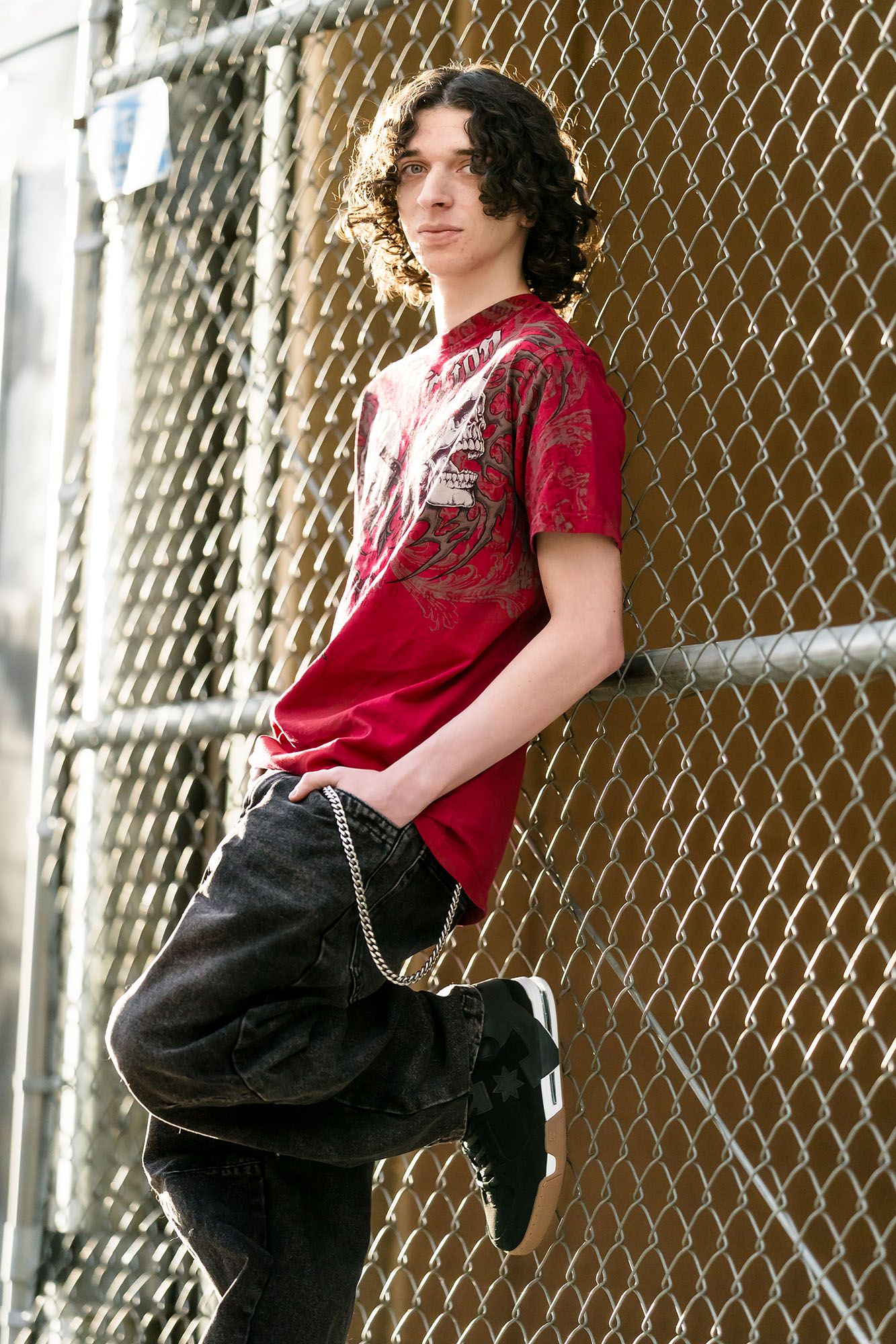 Teen boy with curly hair in red graphic tee leans against chain-link fence in urban setting.