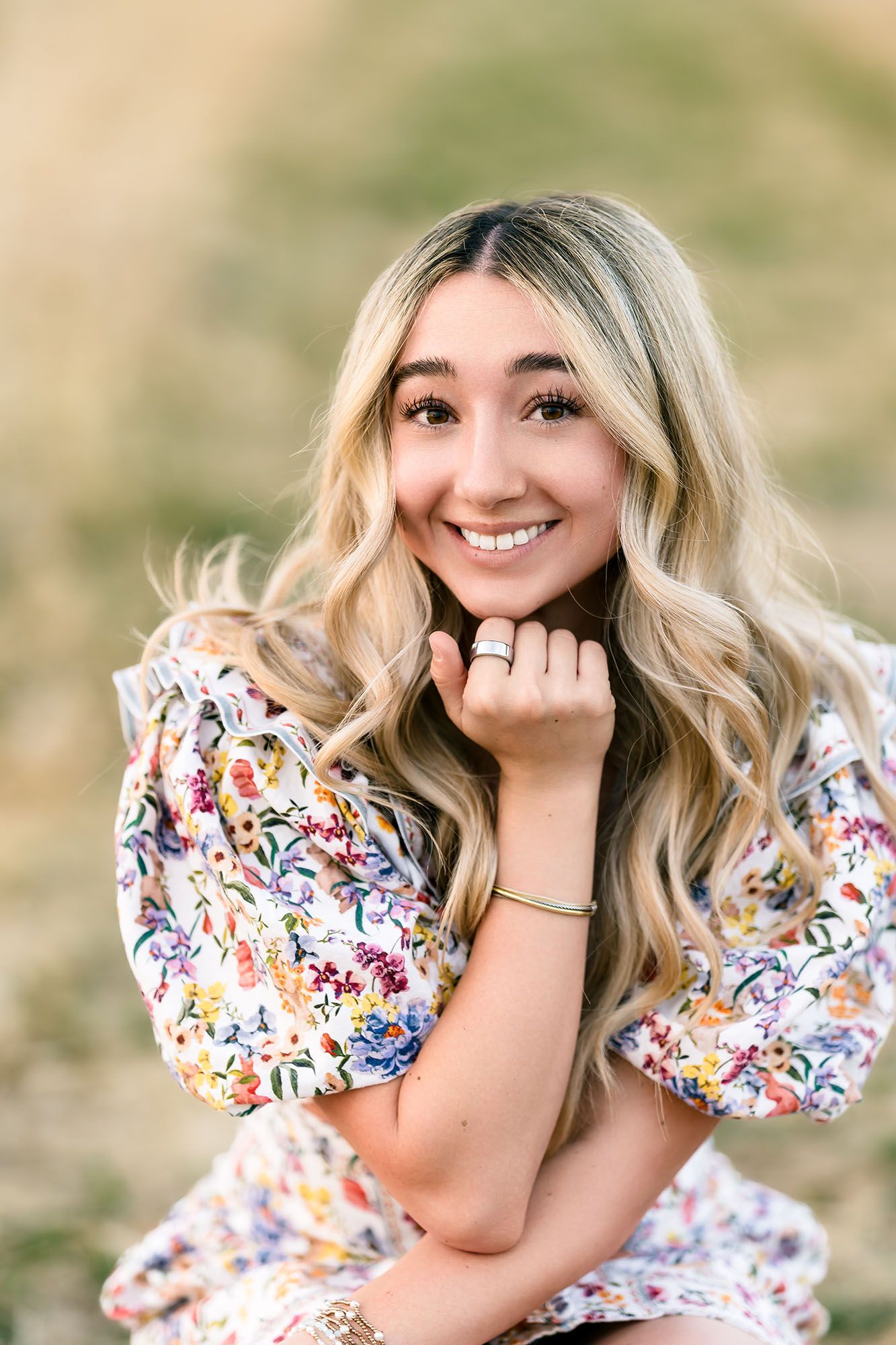 Blonde teenage girl in floral dress smiling warmly while posing outdoors for senior portraits.