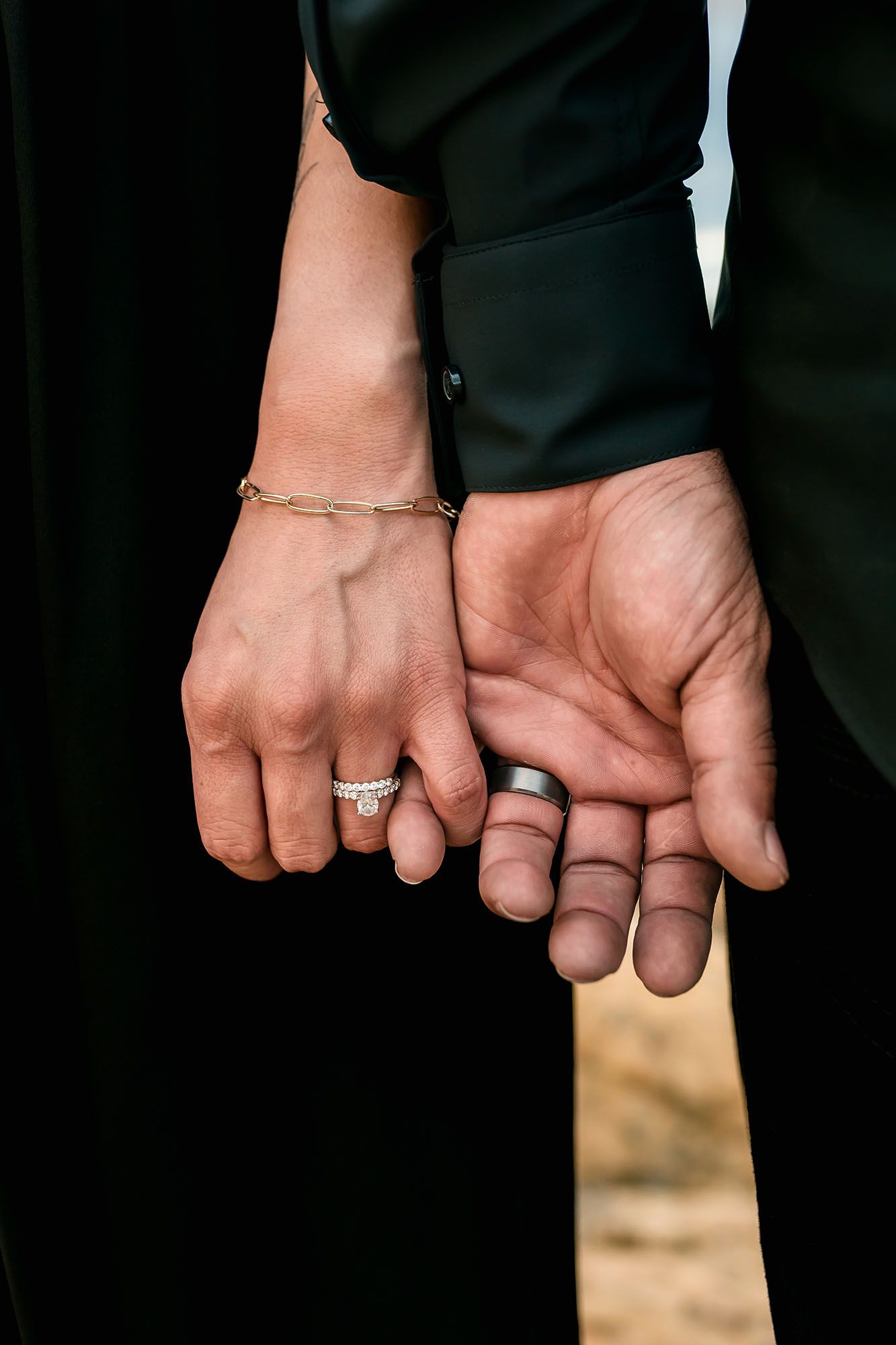 Close-up of couple holding hands showing diamond engagement ring, wedding band, and gold bracelet.