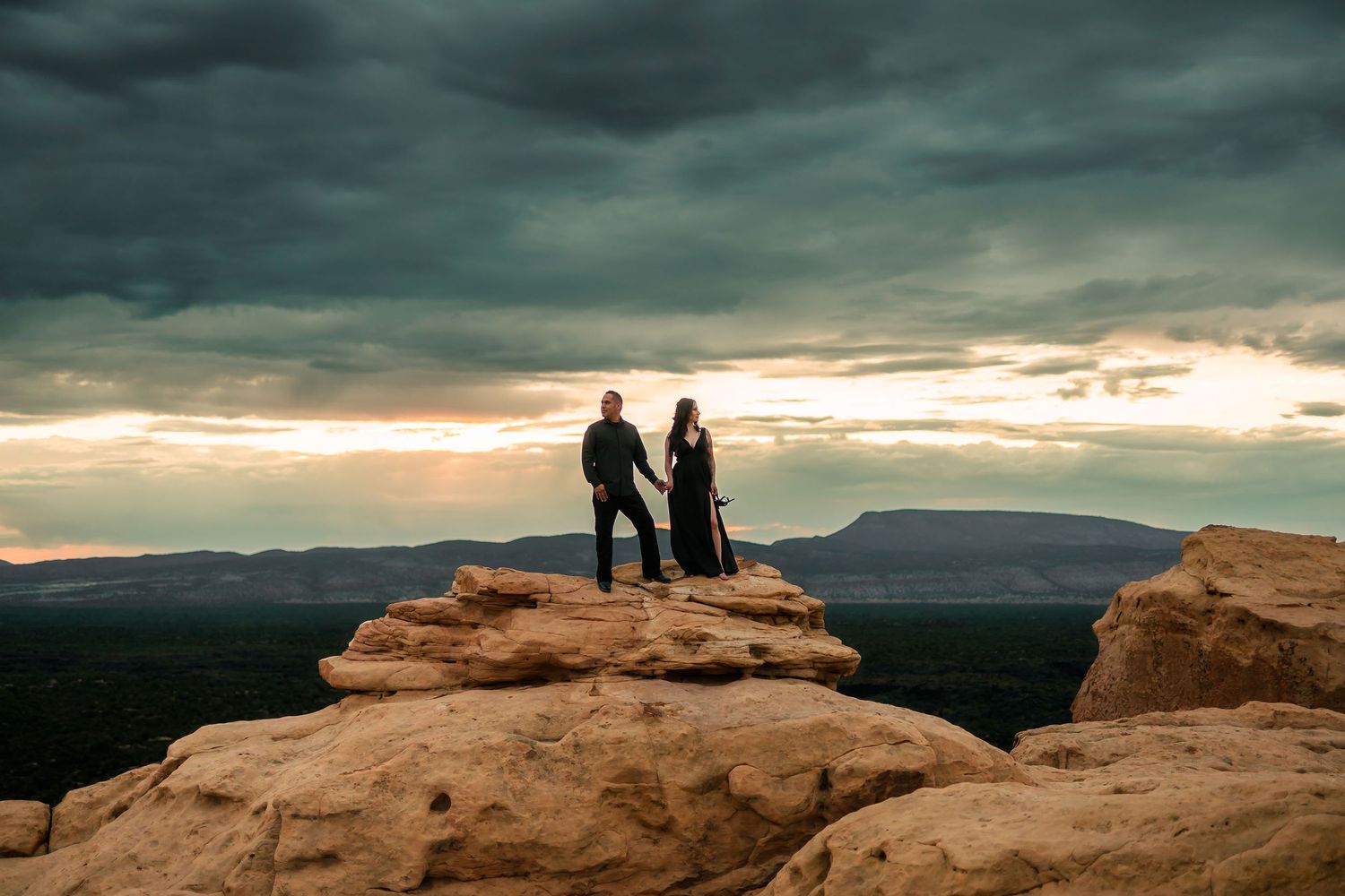Couple standing hand in hand on desert rock formation under dramatic stormy sky.