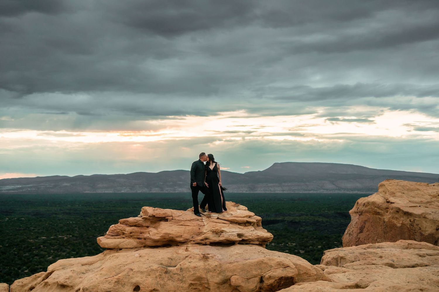 Romantic couple embracing on a sandstone cliff under dramatic stormy skies with mesa views.