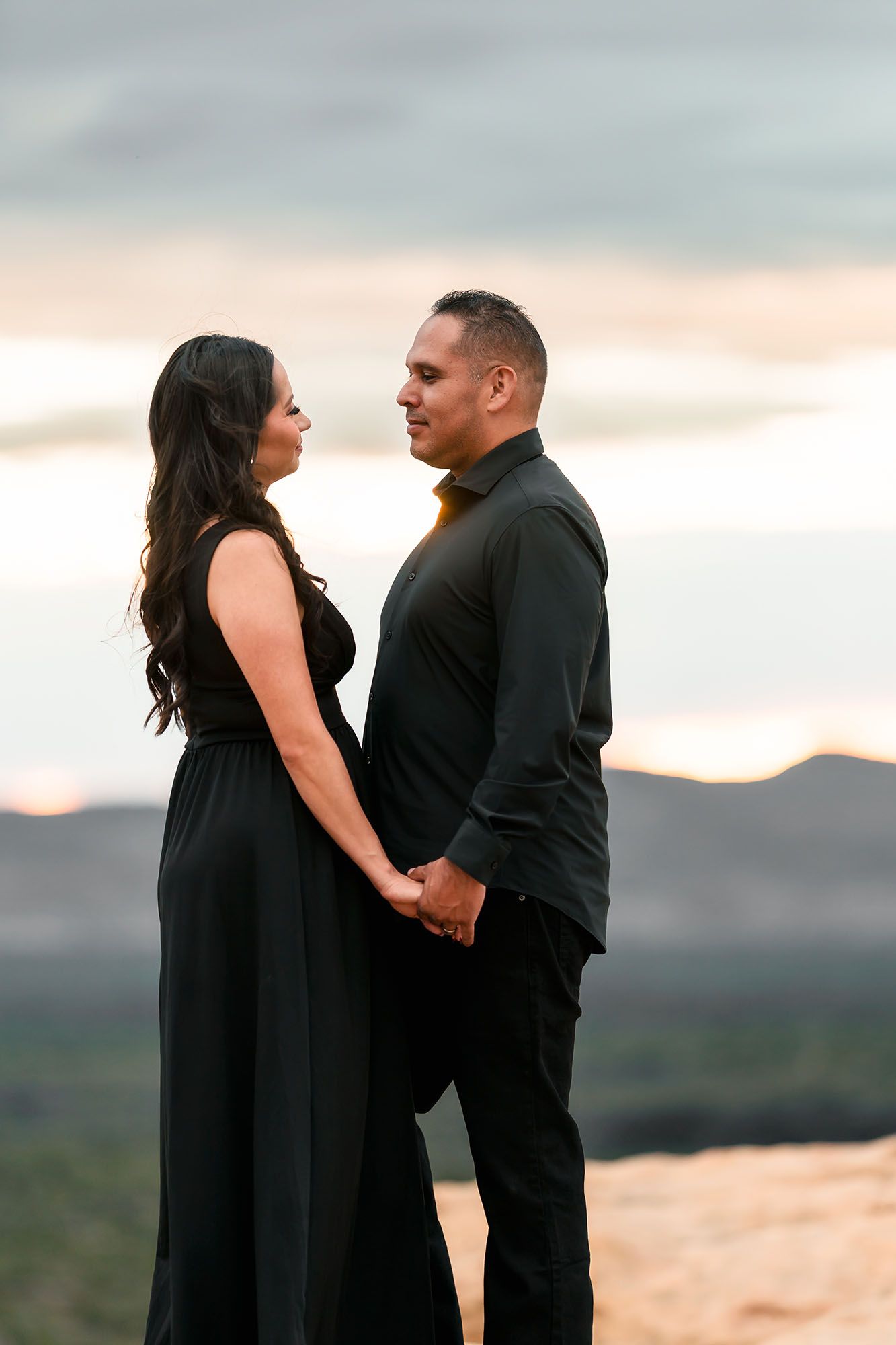 Engaged couple in all-black outfits holding hands at sunset overlooking a scenic desert valley.