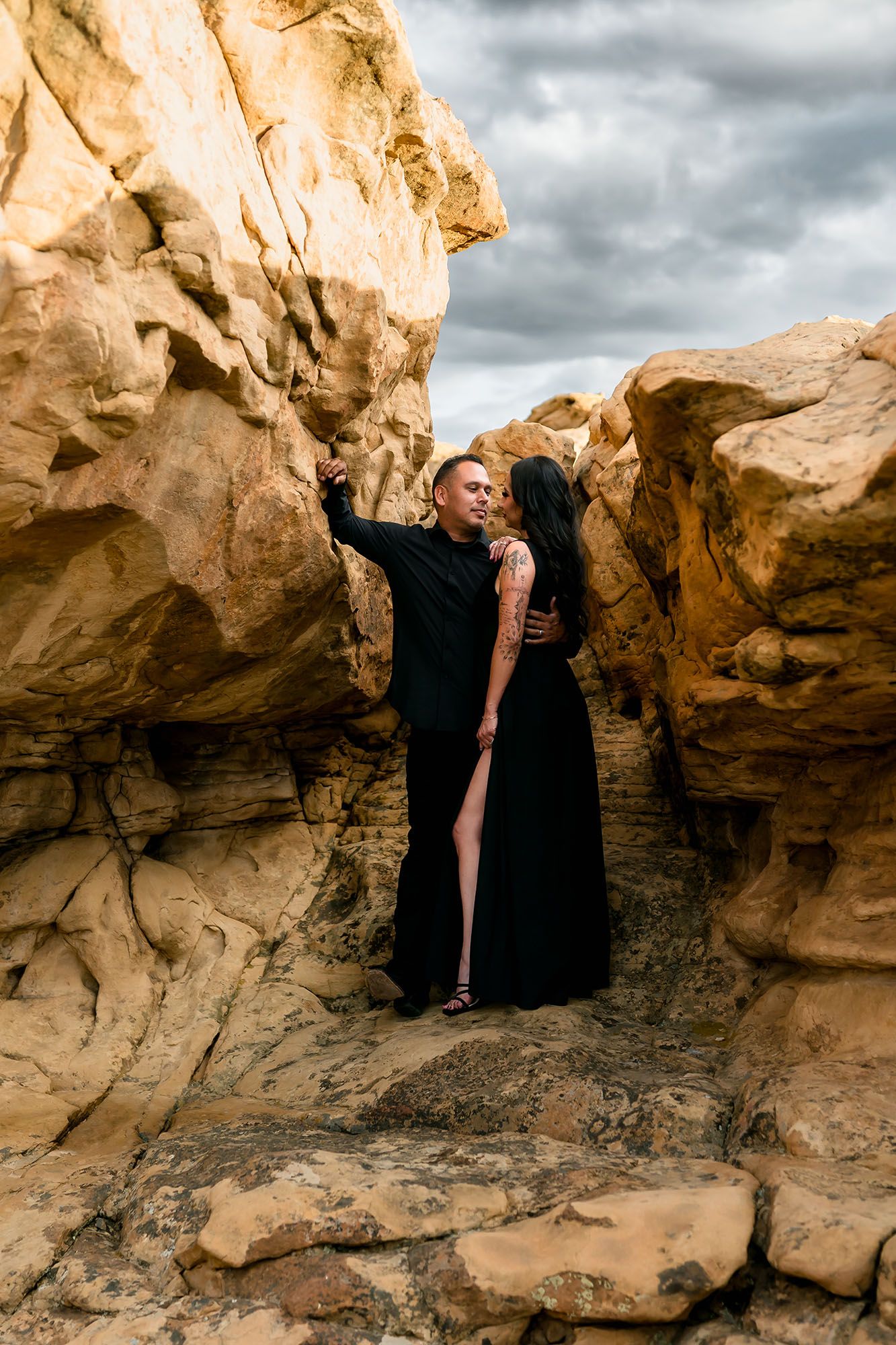 Couple in black outfits posing romantically among golden sandstone rock formations under dramatic cloudy skies.