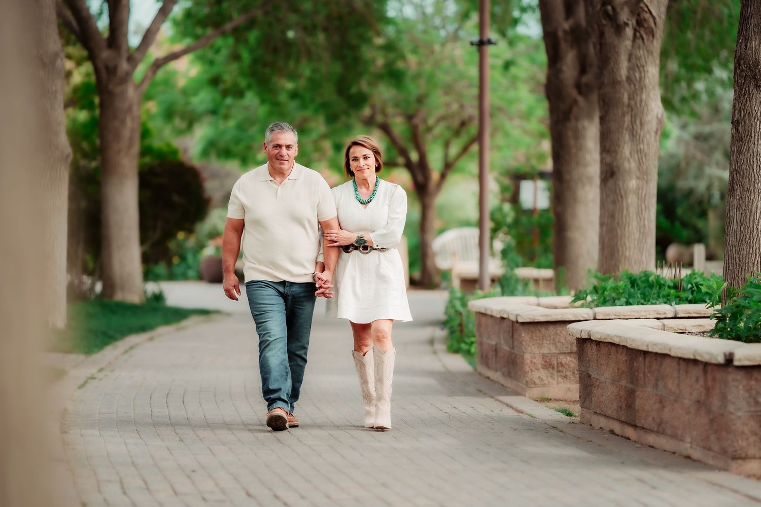 A couple walks together on a tree-lined pathway through a lush green park on a sunny day.