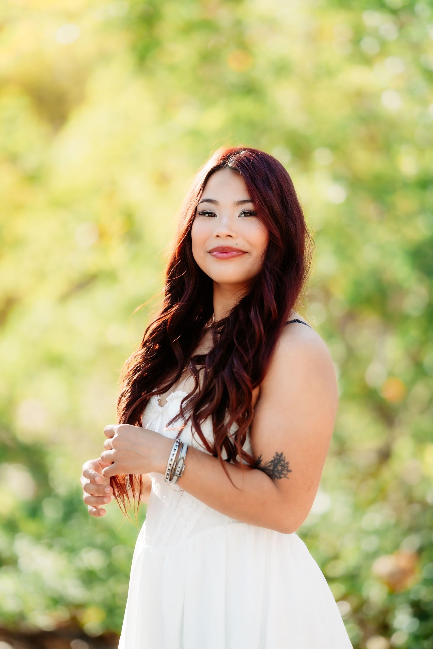 A person in a white dress poses outdoors against a soft bokeh background of green and yellow foliage.